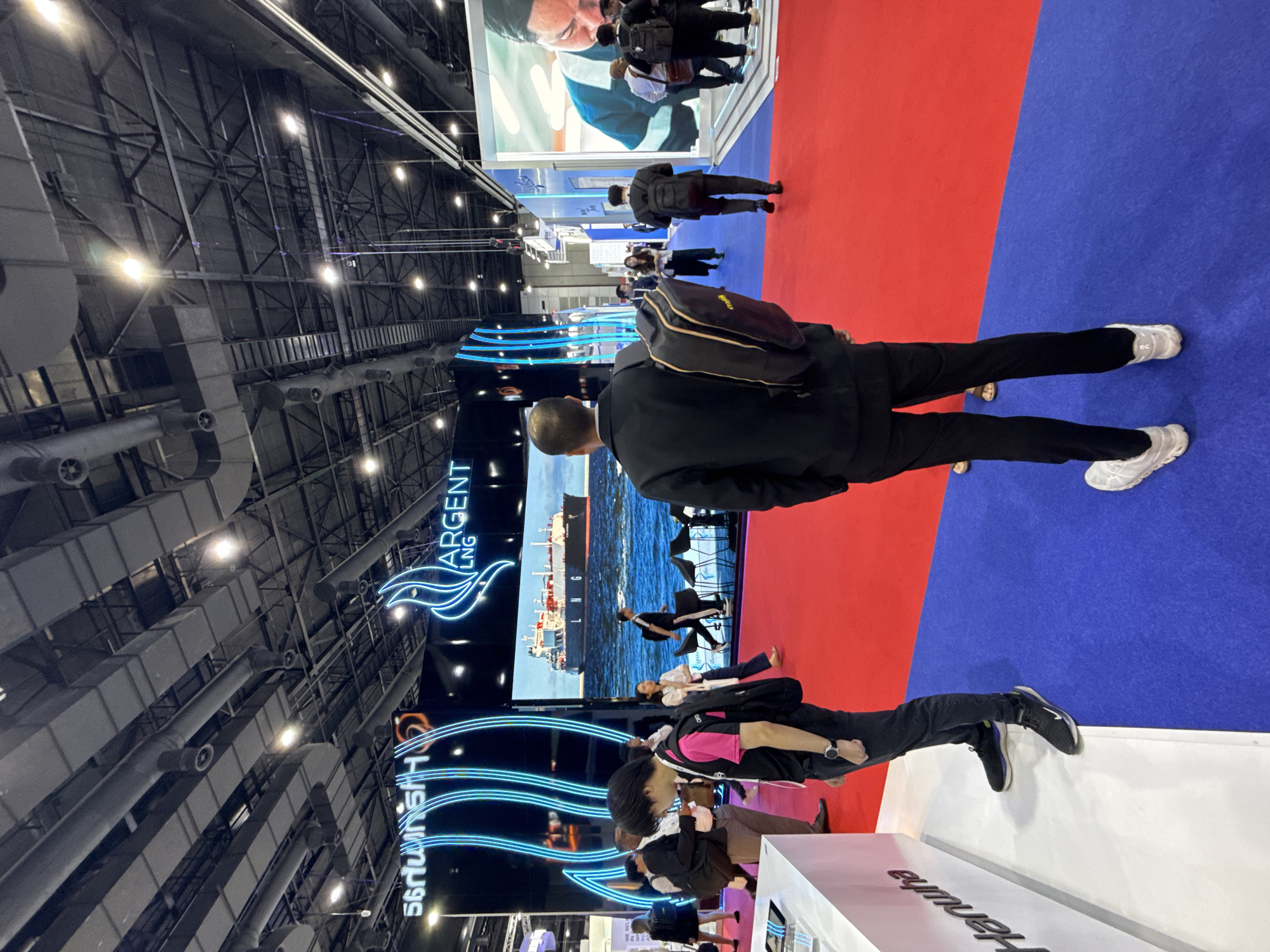 The image depicts an indoor exhibition hall, possibly at the IMPACT Exhibition and Convention Center in Bangkok, Thailand, given the scale and style of the venue. The scene is dominated by a large red carpet leading into the hall, bordered by a blue carpet on the right. Booths with prominent branding and displays are visible.
In the foreground, a person wearing a black suit and a black backpack is standing on the blue carpet, facing away from the camera. To the left, another person in a pink and black shirt and black pants, also with a backpack, is walking on the red carpet. Other attendees, dressed in casual and business attire, are seen browsing the exhibition.
A large digital screen displays a large cargo ship at sea, with "ARGENT LNG" branding above it. Another screen on the left shows illuminated neon-like blue lines and text. Various exhibition stands, lighting fixtures, and ceiling structures are characteristic of a trade show environment. The overall atmosphere suggests a busy event, possibly related to the energy or shipping industry.