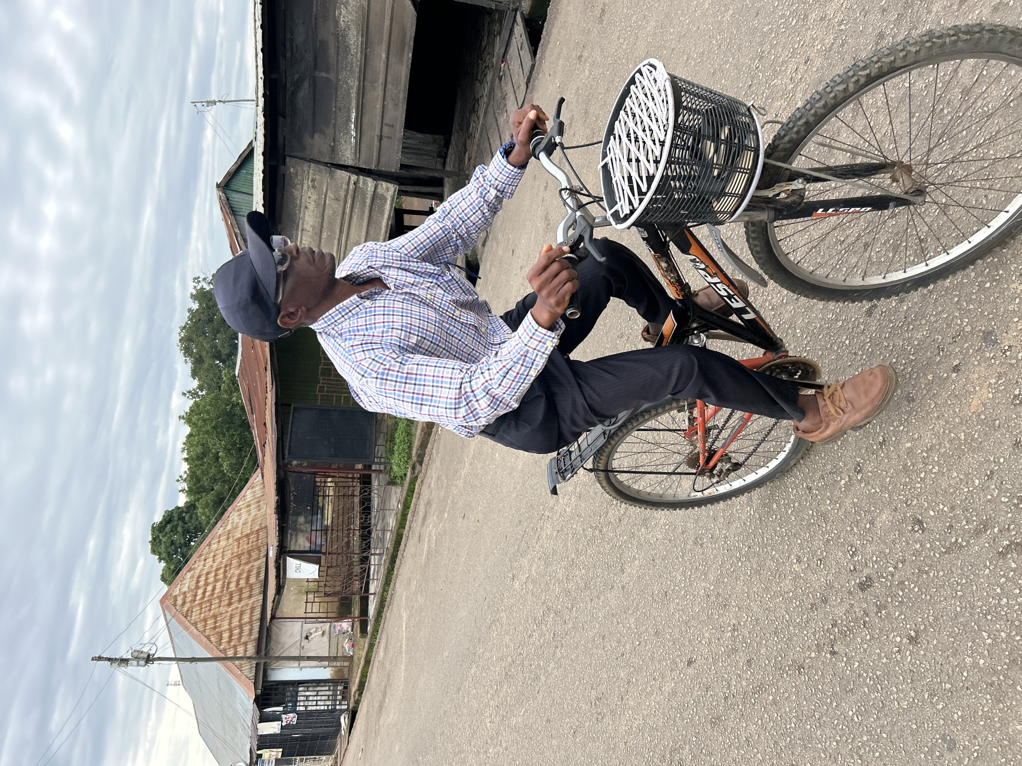 A man wearing a blue and red checkered shirt, black trousers, brown shoes, a dark baseball cap, and glasses rides a bicycle with a black basket on the front. He is on a paved road in what appears to be a semi-urban area. In the background, there are several buildings with corrugated metal roofs, some appearing dilapidated. There are also power lines and utility poles visible. The sky is overcast. The scene is consistent with a street view in Jos, Nigeria, characterized by its architecture and infrastructure.