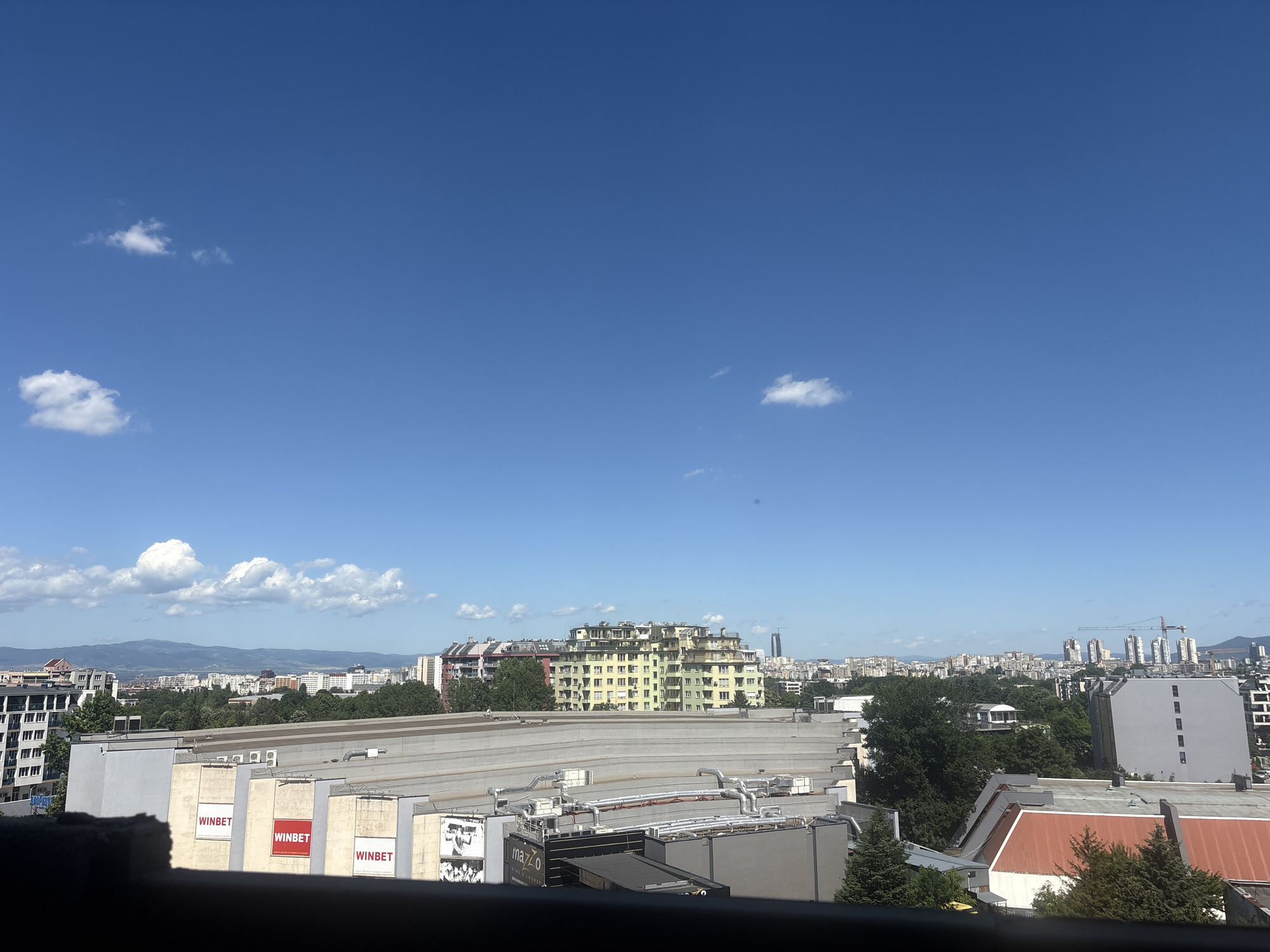 This is an outdoor, eye-level, daytime, horizontal, full shot of a cityscape in Sofia, Bulgaria. The scene is dominated by a bright blue sky with a few scattered white clouds. In the foreground, a large, modern, gray building with a sloped roof is prominent, featuring visible ventilation systems. Several sections of this building display "WINBET" advertisements. To the left and right, other buildings of varying heights and architectural styles are visible, including a tall, light green apartment building with a unique stepped design in the mid-ground. The background shows a sprawling urban landscape with numerous buildings, trees, and distant mountains under the clear sky. Construction cranes are visible in the far distance, indicating ongoing development. No people are clearly discernible in the image. The overall impression is of a sunny day in a densely populated urban area.