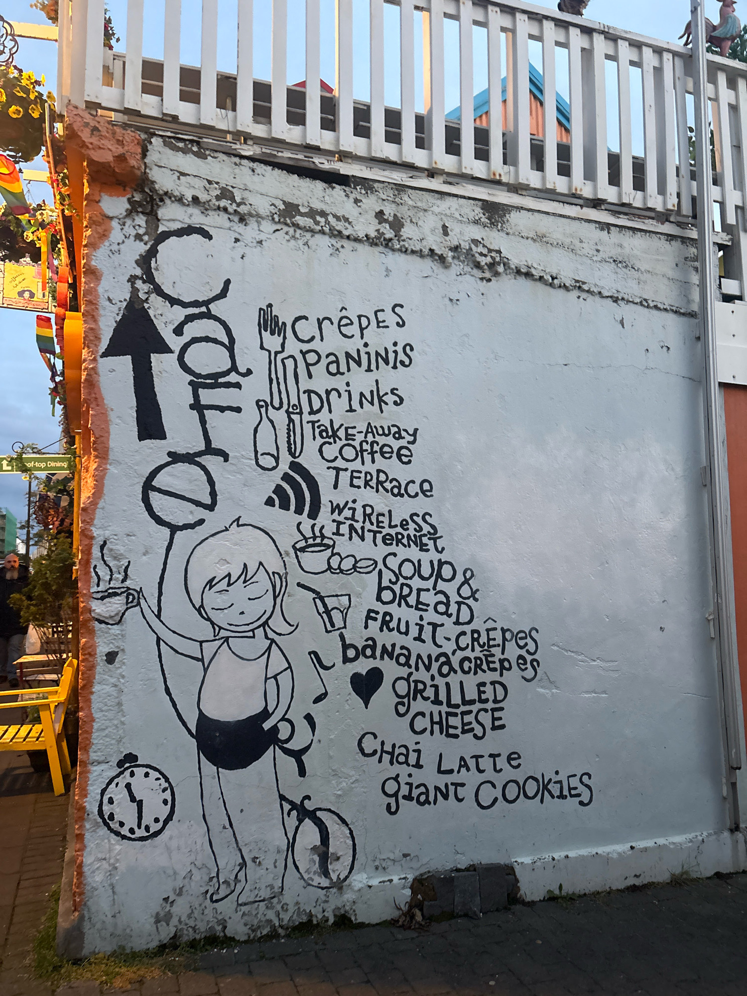 A black and white mural on a concrete wall in Reykjavik, Iceland, advertises a cafe's offerings. The mural features a stylized illustration of a person holding a steaming cup, with various food and drink items listed in hand-drawn script. These include "Crêpes," "Paninis," "Drinks," "Take-Away Coffee," "Terrace," "Wireless Internet," "Soup & Bread," "Fruit-Crêpes," "Banana Crêpes," "Grilled Cheese," "Chai Latte," and "Giant Cookies." An arrow points upwards, and a clock is drawn on the lower left. The wall shows signs of age and wear. In the background, a white railing overlooks a building with a blue roof. To the left of the mural, a yellow bench is visible, along with hanging plants and rainbow-colored banners. A street sign with "L'-top Dining" is also present.
