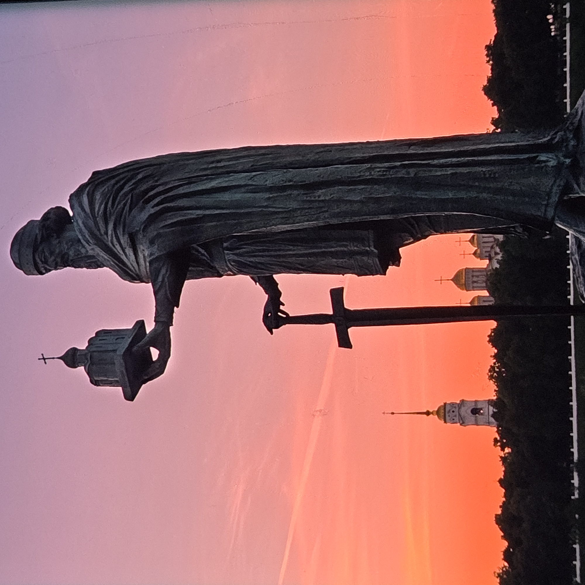 The image depicts a full-body bronze statue of Prince Vladimir the Great, holding a model of the Dormition Cathedral in his right hand and a sword in his left. The statue stands against a vibrant sunset sky that transitions from pink to orange. In the background, silhouetted trees line the horizon, with several golden-domed churches and a bell tower visible. This scene is located in the city of Vladimir, Russia, specifically on the Prince Vladimir Monument, overlooking the Klyazma River. The monument was erected in 2004.