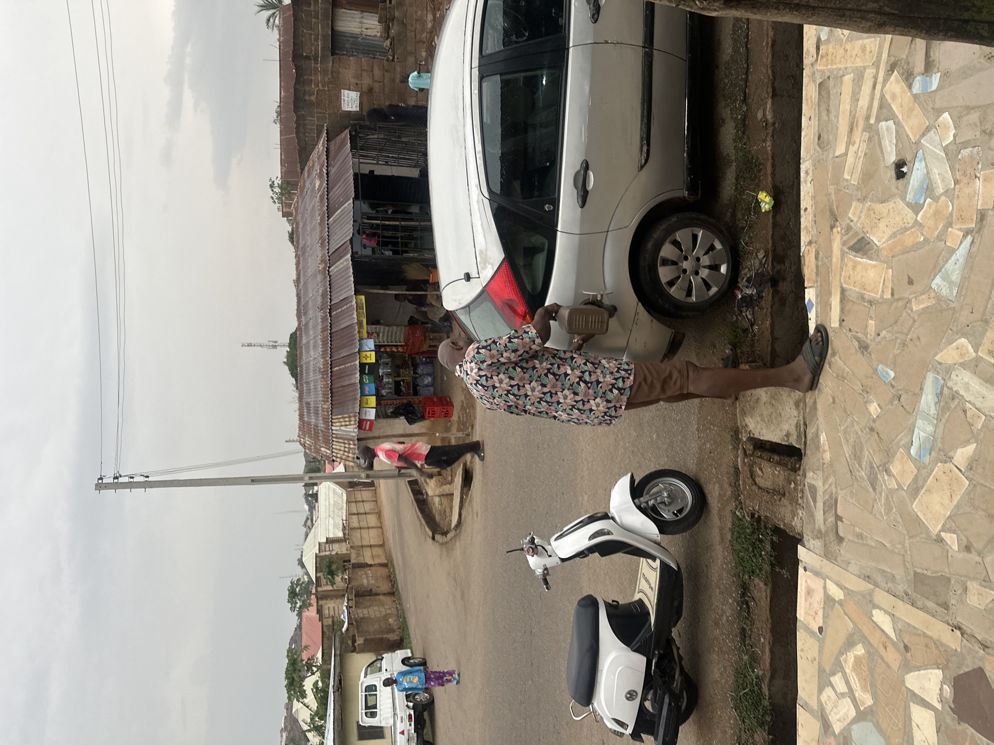 A man in a floral shirt and brown shorts is pouring gasoline from a brown container into a white car parked on the side of a street. A white motor scooter is parked behind the car, and a small store with a corrugated metal roof stands in the background. Another person is sitting near the store. The street slopes downwards and features a white truck and two figures in the distance. The scene appears to be set in a residential neighborhood with additional buildings and a utility pole visible in the background. The scene shows the man refilling fuel, and this might be happening in the city of Jos in Nigeria. The street and sidewalk surface feature different materials and finishes.