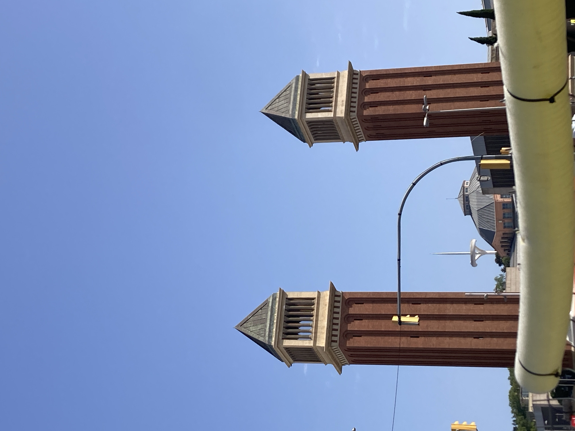 The image displays two Venetian-style towers made of red brick with a lighter stone top section and a dark pyramidal roof, set against a clear blue sky. These towers are located in Barcelona, Spain, at the base of Montjuïc hill, flanking the main entrance to the former Fira de Barcelona exhibition grounds. In the middle ground, a curved traffic light arm spans between the towers, and a traffic light with a yellow signal is visible on the left tower. Beyond the towers, the unique spire of the Torre de Comunicaciones de Montjuïc and the distinctive domed roof of the Palau Nacional are discernible. The foreground is partially obscured by a thick, bright yellow cylindrical object, possibly part of a vehicle or street furniture, secured with black zip ties. The scene suggests a daytime outdoor environment.