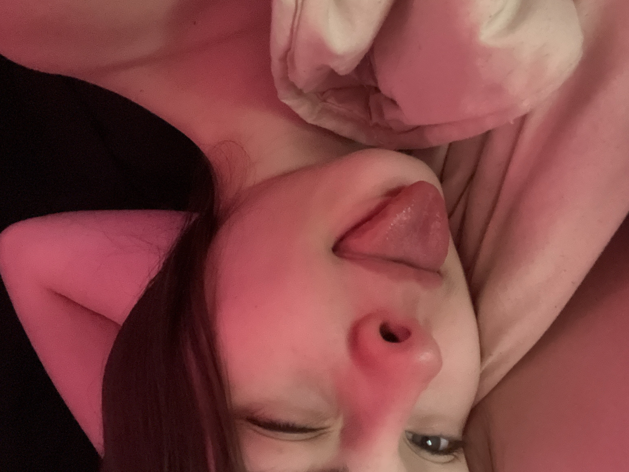 A close-up, angled photograph features a fair-skinned individual with dark hair and partially closed eyes, sticking their tongue out. The person is in a recumbent position, possibly in a bed, with bedding and a dark garment visible in the background. The lighting is predominantly pink, casting a soft glow on the subject and surrounding objects. Location information is not discernable from the image and cannot be confirmed.