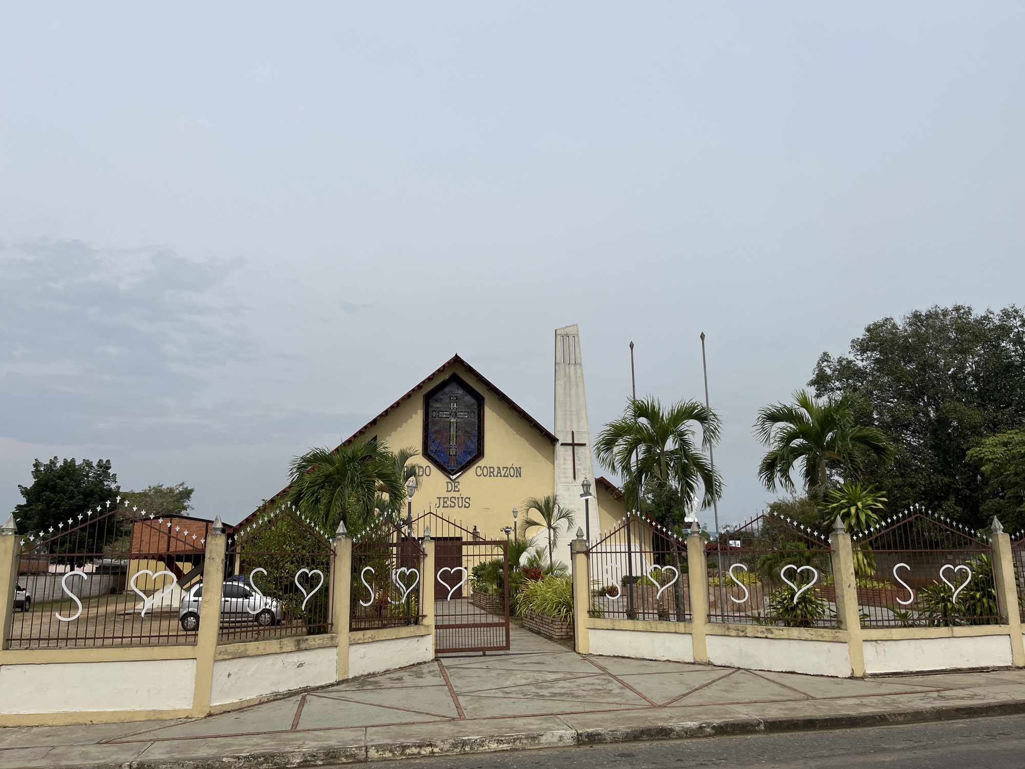 The image captures the "Sagrado Corazón de Jesús" church located in Tinaquillo, Venezuela. The church is characterized by its light yellow facade, a prominent stained glass window featuring a cross at the apex of the roof, and a slender, modernist bell tower to the right. A wrought-iron fence with heart and "S" shaped decorations runs along the front of the property, with a gate providing access to the church. Palm trees flank the entrance, adding a tropical element to the scene. The sky is overcast, and a section of the street is visible in the foreground. A few cars are visible parked inside the property.