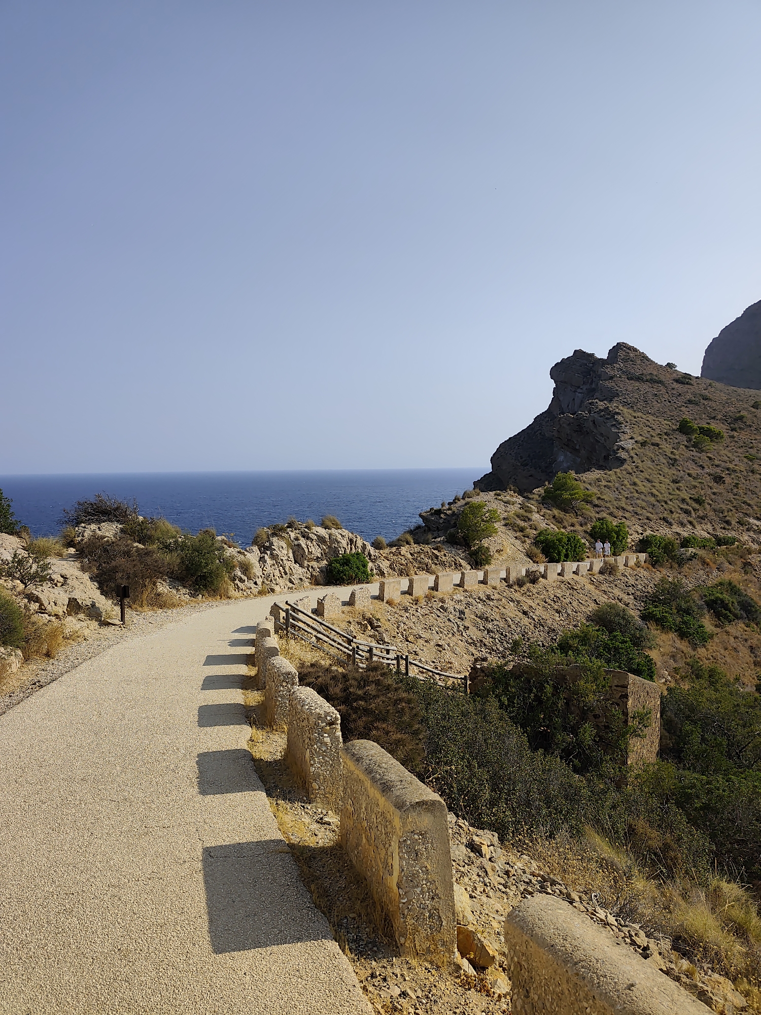 The image depicts a coastal road winding along a rocky, arid hillside. The road is paved with light-colored gravel and is bordered by a low stone wall topped with concrete pillars. Beyond the wall, a wooden fence runs along the edge of the road. The sea, a deep blue, stretches to the horizon under a clear, pale blue sky. Sparse, dry vegetation and shrubs grow on the hillside, interspersed with larger rocks and some trees. In the distance, two people in white clothing are walking along the road, near a rocky outcrop. The overall impression is of a scenic, somewhat remote coastal location, likely in a Mediterranean climate. This scene is characteristic of the coastal paths found in the vicinity of L'Alfàs del Pi, Spain, known for its rugged coastline and sea views.