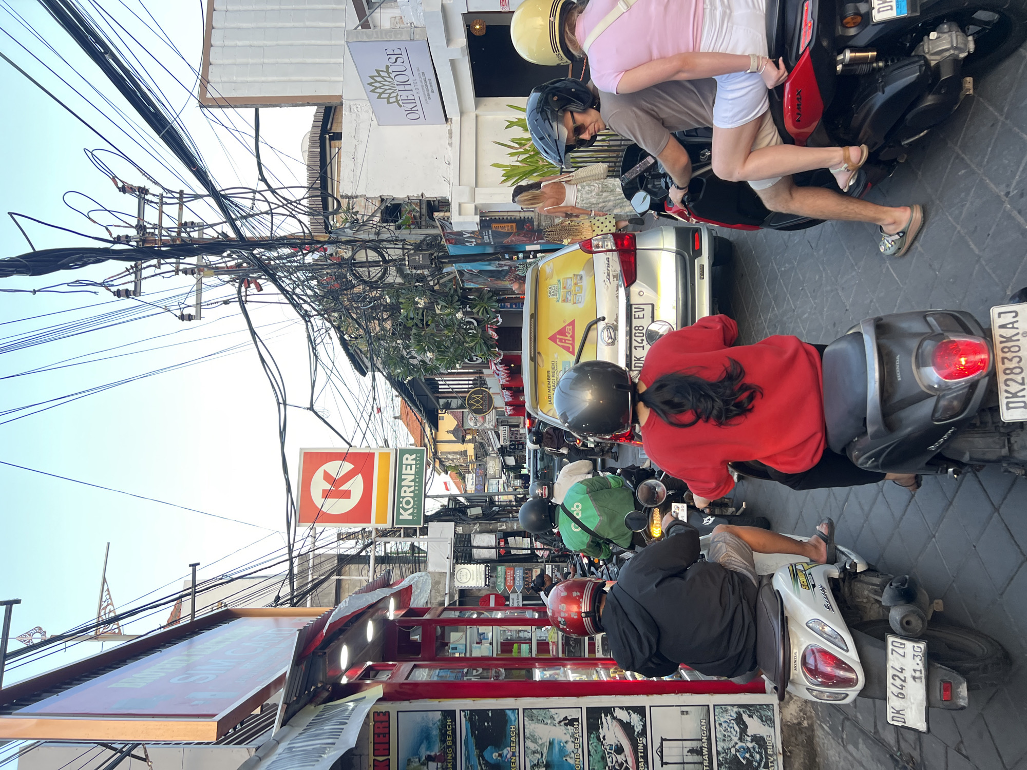 This is a street scene in Kuta, Indonesia, with heavy traffic congestion. A group of people on scooters are the main subjects, navigating through the crowded street. The atmosphere is bustling, with shops lining both sides of the road and a tangle of electrical wires overhead.
The scene is shot from behind a person on a white scooter, who is wearing a black jacket and a red helmet. To their right, another person on a red scooter is visible, wearing a red shirt and a black helmet. In front, a silver car is stopped in traffic, with several people walking on the sidewalk behind it. Further down the street, more scooters and cars are visible, indicating a busy urban environment.
The shops on the left side of the street display various advertisements and products, suggesting a commercial district. The buildings are a mix of traditional and modern architecture. The electrical wires are prominently featured, creating a complex network above the street. The overall impression is one of a vibrant, densely populated area experiencing a typical day of traffic and commerce.