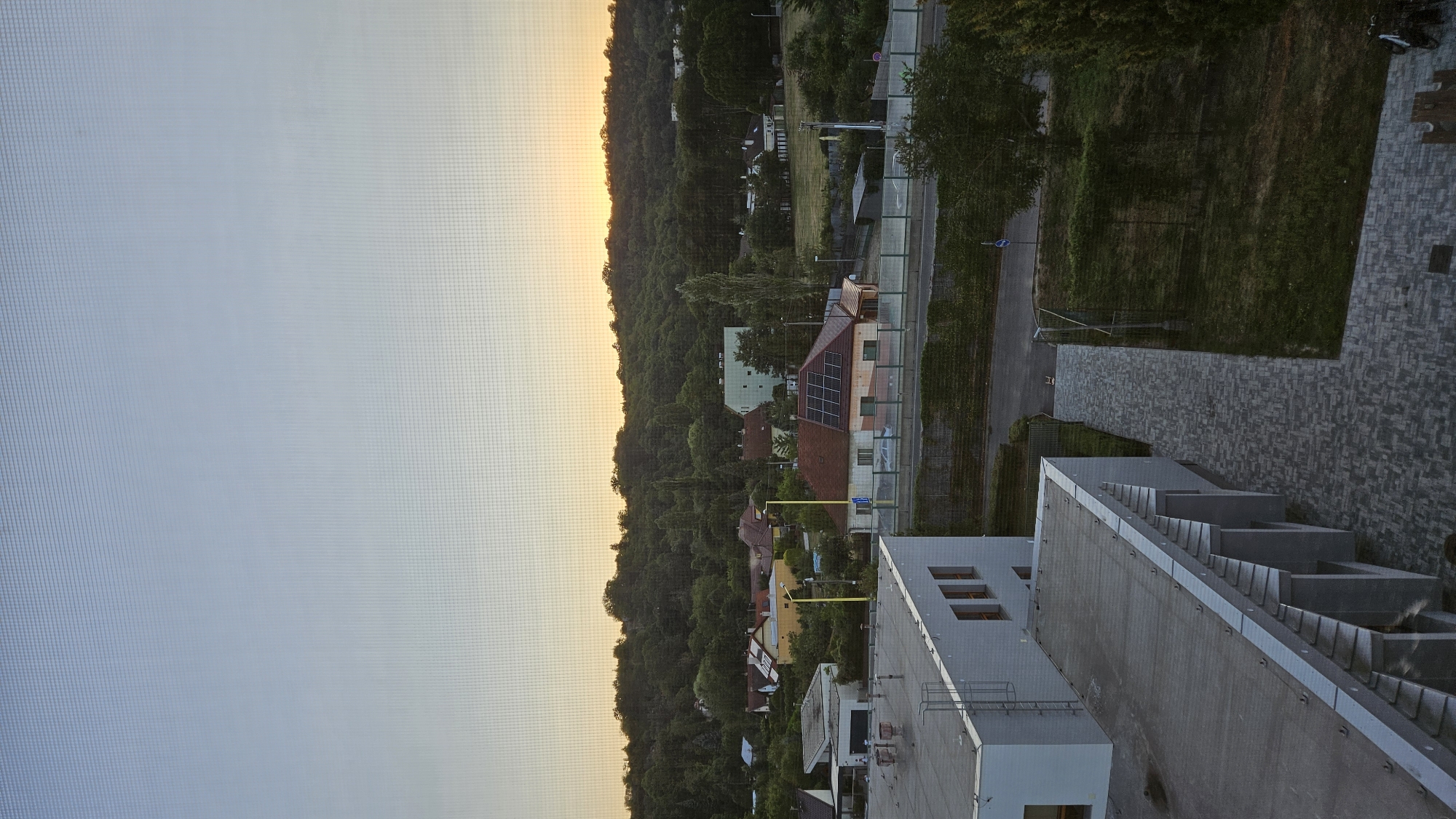 The image captures a twilight view over a suburban area, likely from an elevated position, with a hazy sky transitioning from pale blue to a soft orange glow near the horizon. In the foreground, the modern architecture of a flat-roofed building with light-colored walls and visible exterior conduits is prominent. Adjacent to it is a paved walkway with a dark, patterned surface.
Beyond the foreground building, a street is visible, bordered by green hedging and a chain-link fence. Several houses with reddish-brown roofs and some with solar panels are scattered across the mid-ground, nestled amongst lush green trees. The landscape is dominated by a densely wooded hill that forms the backdrop, with the setting sun casting a warm light on its upper edge.
The presence of a few parked cars, streetlights, and what appears to be a sports field or open grassy area suggests a residential and possibly institutional setting. The overall impression is one of a quiet, developing urban fringe as dusk settles. Given the request to locate it in Prešov, Slovakia, this scene could represent a view from a building in the Sekčov housing estate looking towards the surrounding hills and the suburban development beyond. There are no individuals visible in the frame.