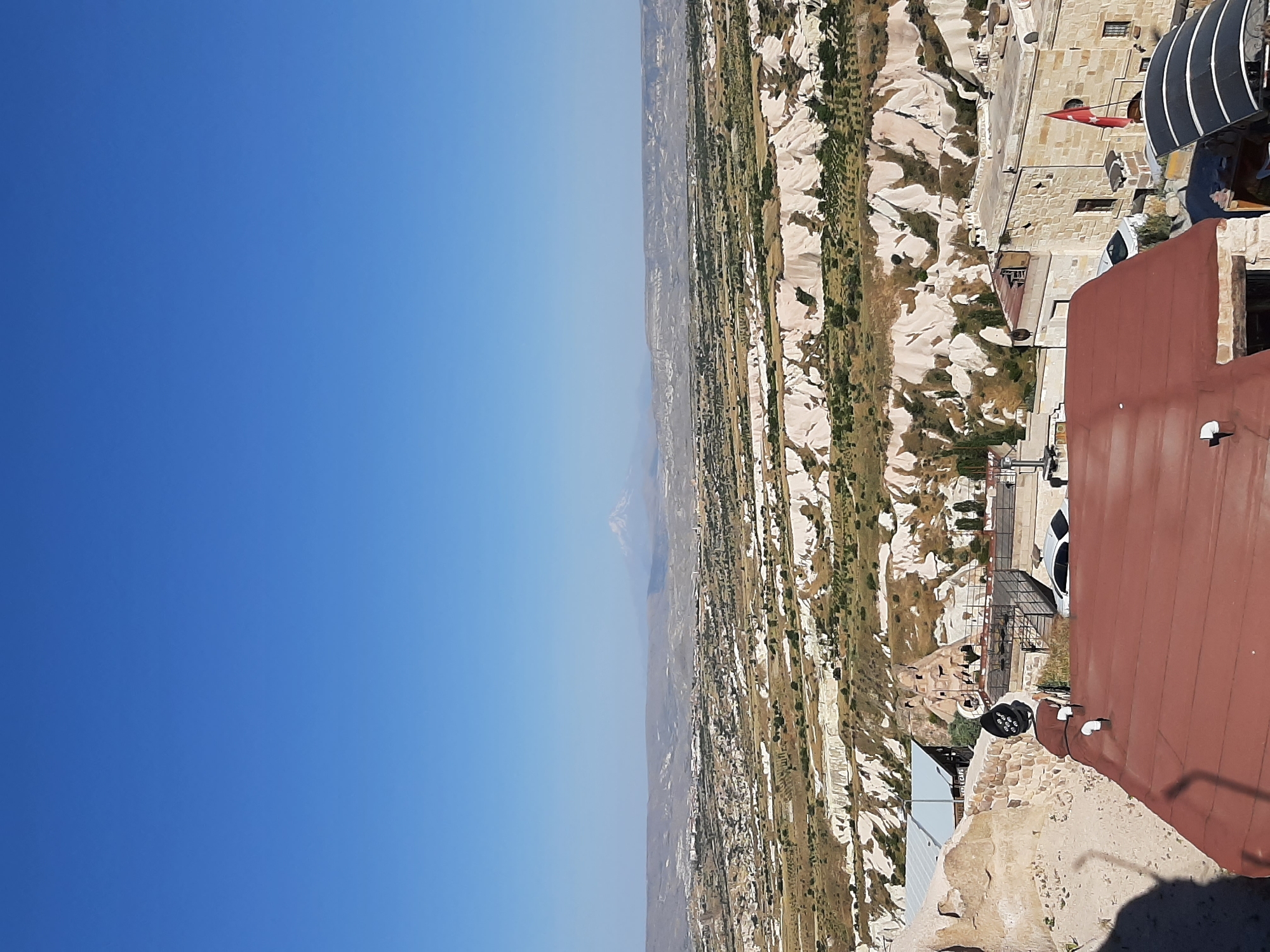 This image captures a panoramic view of Uçhisar Beldesi, Turkey, showcasing its unique geological formations and urban landscape under a clear blue sky. In the foreground, a rust-colored roof dominates the lower part of the frame. To the left, a stone wall with a rough texture is visible, partially obscuring a light-colored structure behind it.
In the middle ground, several buildings are scattered across the terrain. One building has a light-colored stone facade and a Turkish flag flying from it. Adjacent to this, another building features a darker, possibly metal roof, and a sign that appears to read "i cafe". Further back, unique rock formations, characteristic of Cappadocia, rise from the landscape, some with openings that suggest they might have been inhabited or modified. A white car is parked in front of one of the buildings.
The background reveals a vast, undulating landscape characterized by soft, layered rock formations in shades of white and tan, interspersed with patches of green vegetation. In the far distance, a majestic snow-capped mountain, likely Mount Erciyes, stands prominently against the horizon, its peak sharply defined against the hazy sky. The overall scene conveys a sense of arid beauty and the distinctive architectural style of the region.