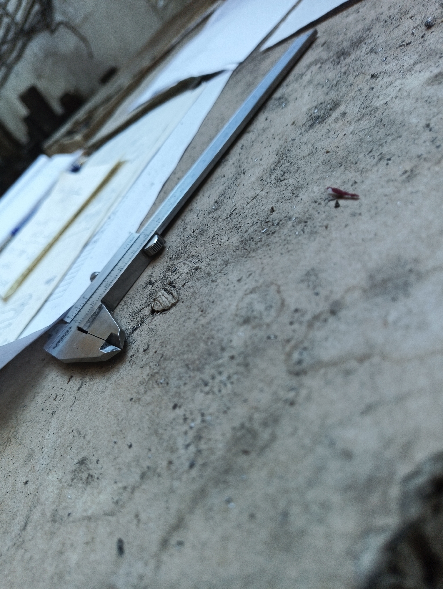 A close-up, eye-level shot shows a metal caliper placed diagonally on a rough, grey concrete surface. The caliper is silver with black markings and is positioned over several white sheets of paper. The background is out of focus, showing a blurry, dimly lit area with some vertical structures. A tiny, reddish object, possibly an insect or a piece of debris, is visible on the concrete to the right of the caliper. The overall scene suggests an outdoor or workshop environment. The city of Karnobat, Bulgaria, is not visible in this image.