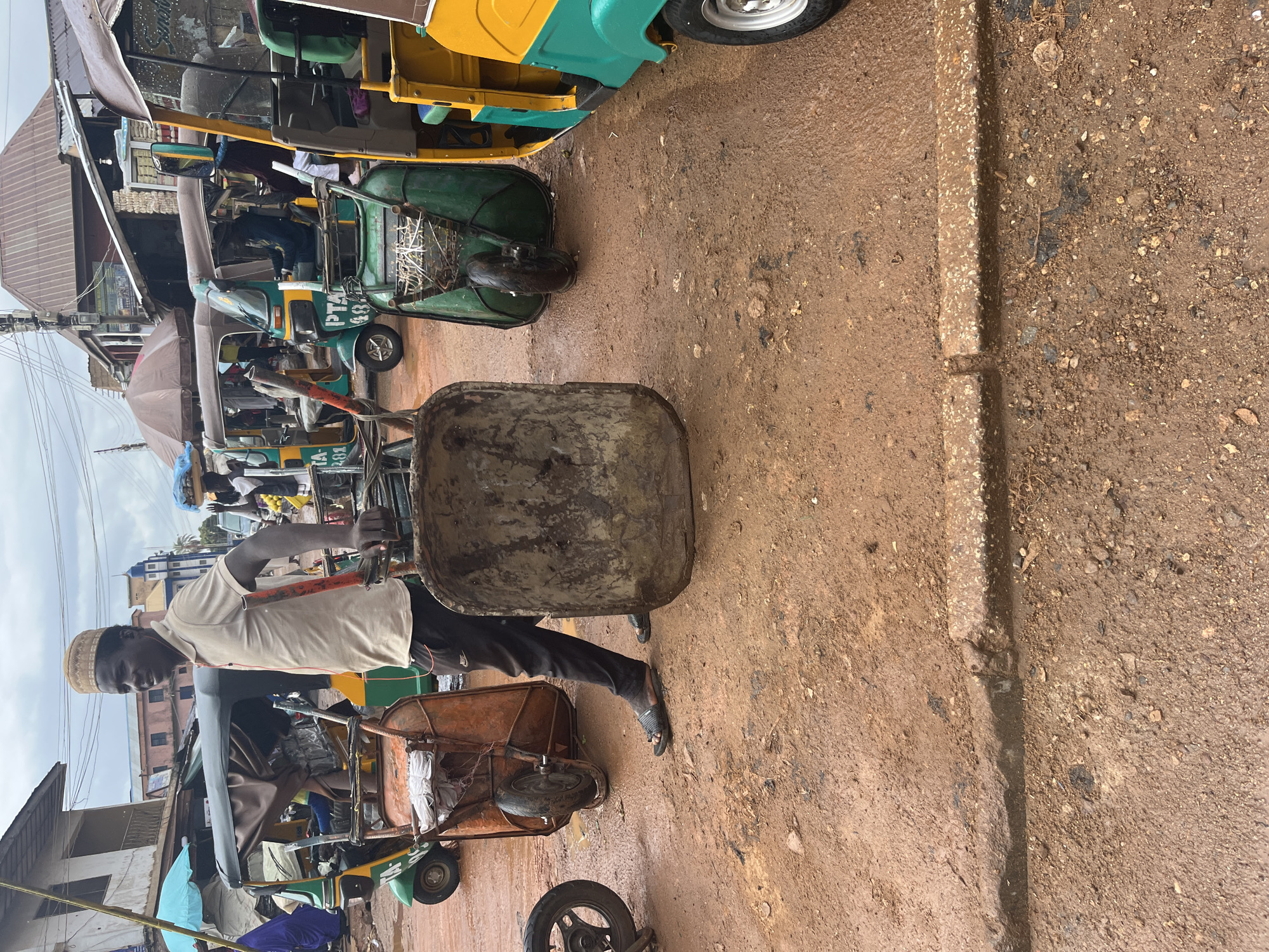 The image depicts a man in a tan polo shirt and dark pants standing on a dirt road, holding the handles of a large, empty wheelbarrow. He is wearing a patterned cap and sandals. To his left, another wheelbarrow, orange and rusty, is positioned with its wheel visible. In the background, several auto-rickshaws, also known as Keke Napeps, are parked. The rickshaws are predominantly green and yellow, with some having canopies. The environment appears to be a marketplace or a busy street in Jos, Nigeria, characterized by the unpaved, muddy ground and the presence of commercial vehicles. The sky is overcast.