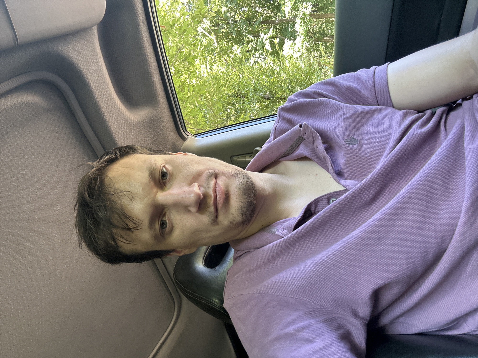 A man is sitting inside a vehicle, with trees visible through the open window. The man is wearing a light purple polo shirt with a small emblem on the left chest. He has light brown hair and a light beard. The interior of the car is visible, including the headliner and a portion of the driver's seat. The scene appears to be outdoors, possibly in a rural or suburban area, given the dense foliage outside the window. No specific location in Shatava, Ukraine can be identified from the image.