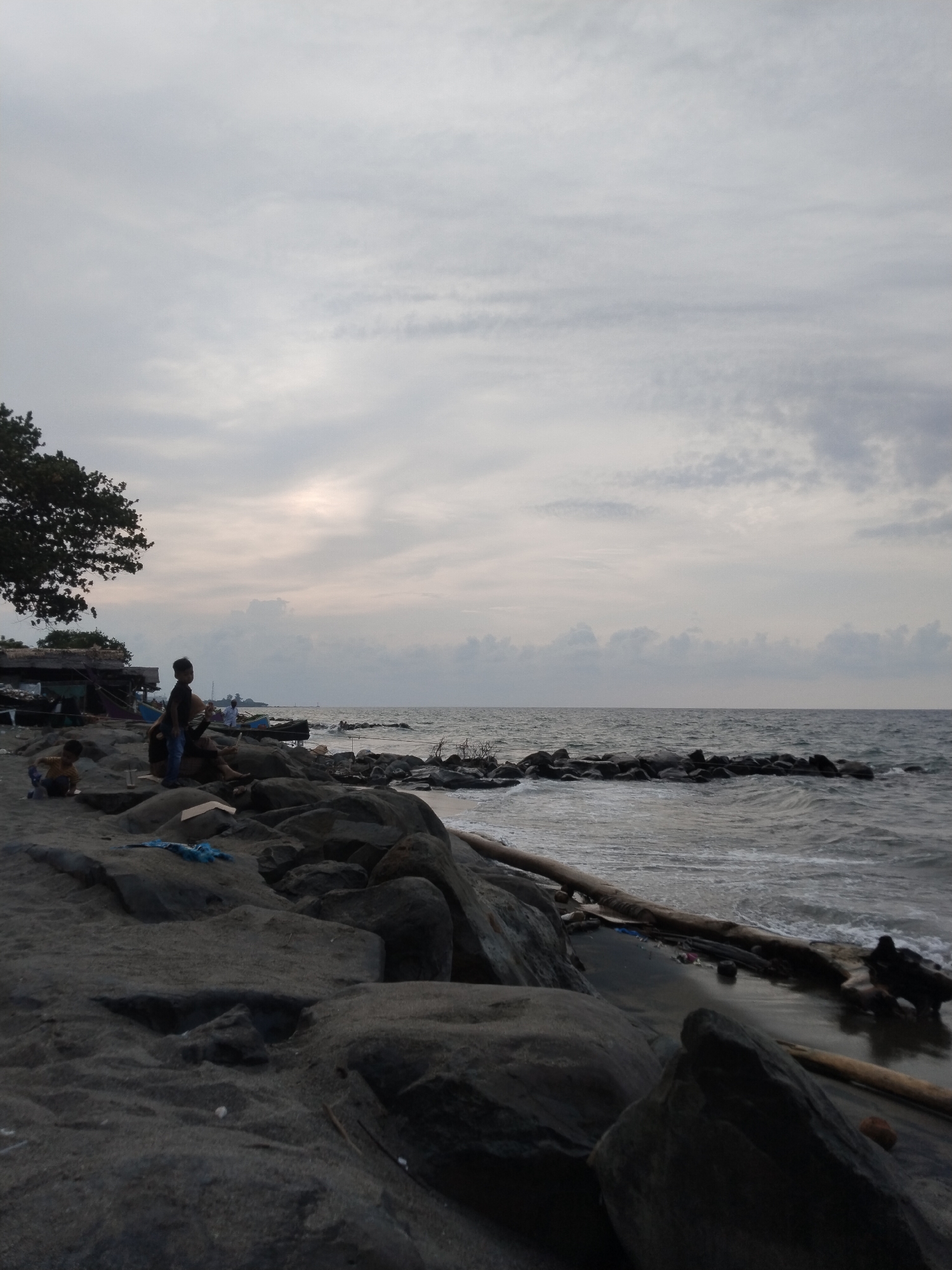 The image shows a beach in Lhokseumawe, Indonesia, under a cloudy sky. In the foreground, large dark rocks and driftwood are scattered on the dark sand. Two children are seen near the rocks on the left of the frame; one is sitting while the other is standing nearby with what appears to be an adult sitting down as well. Fishing boats and a simple shelter are visible in the background. In the sea, a rocky breakwater extends horizontally across the frame. The sea is choppy, with small waves breaking against the rocks.