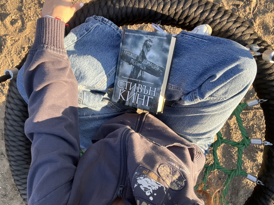 An overhead view captures a person seated on a dark rope nest swing with a green net base. The individual wears light blue denim jeans, a dark grey or navy zippered hoodie with white graphic elements on the left side, and white sneakers. A red thread bracelet is visible on the subject's right wrist. An open book is resting on their lap, identified as "СТИВЪН КИНГ ГРОБИЩЕ ЗА ДОМАШНИ ЛЮБИМЦИ" (Stephen King, Pet Sematary) in Cyrillic script, featuring a dark cover with a prominent image of a cross. Direct sunlight illuminates the scene, creating distinct shadows. The ground beneath the swing appears to be light-colored earth or sand, suggesting an outdoor setting. The scene is located in Maastricht, Netherlands.