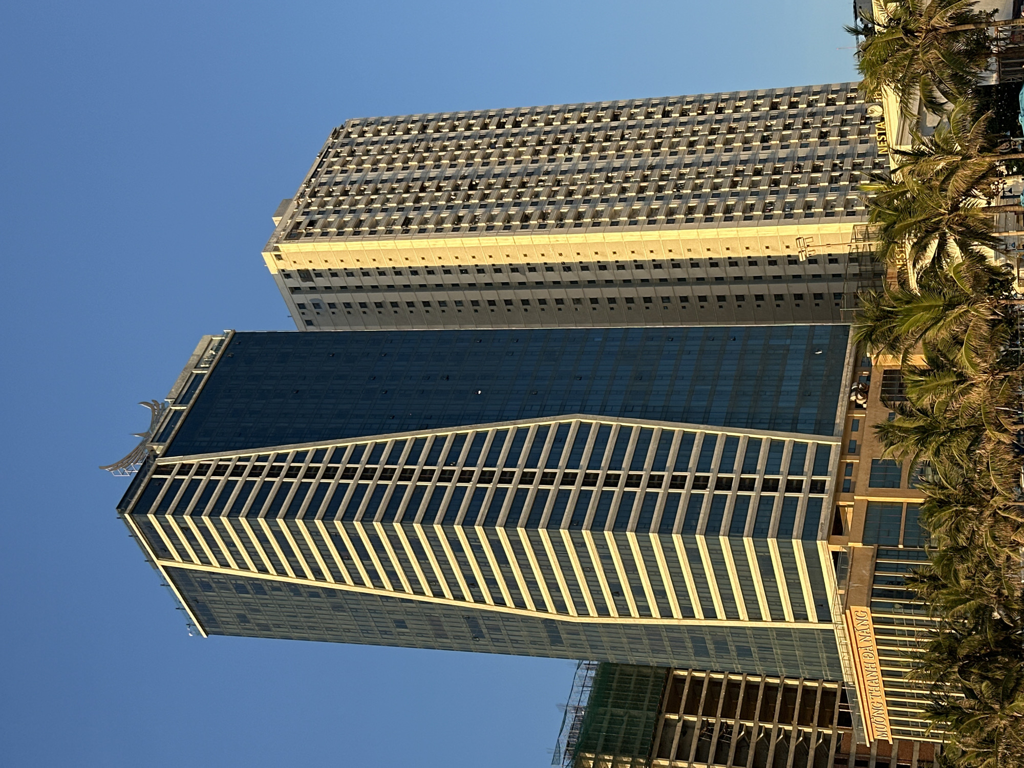 Two prominent skyscrapers dominate the scene against a clear blue sky. The skyscraper on the left is tall and sleek, featuring a distinctive design with angled white stripes alternating with dark glass panels, giving it a stepped appearance. A decorative, curved metallic element adorns the very top of this building. To its right stands a taller, more monolithic skyscraper with a uniform grid of windows and balconies, reflecting the warm sunlight.
In the foreground, a low-rise building is visible, adorned with a sign that reads "MƯỜNG THANH ĐÀ NẴNG" in illuminated lettering. This building is partially obscured by a line of palm trees, their fronds creating a natural fringe across the bottom of the image. Further to the right, another building with "NESTA" prominently displayed is partially visible behind the palm trees. The overall impression is one of a modern urban environment, likely a coastal city, with significant architectural development.