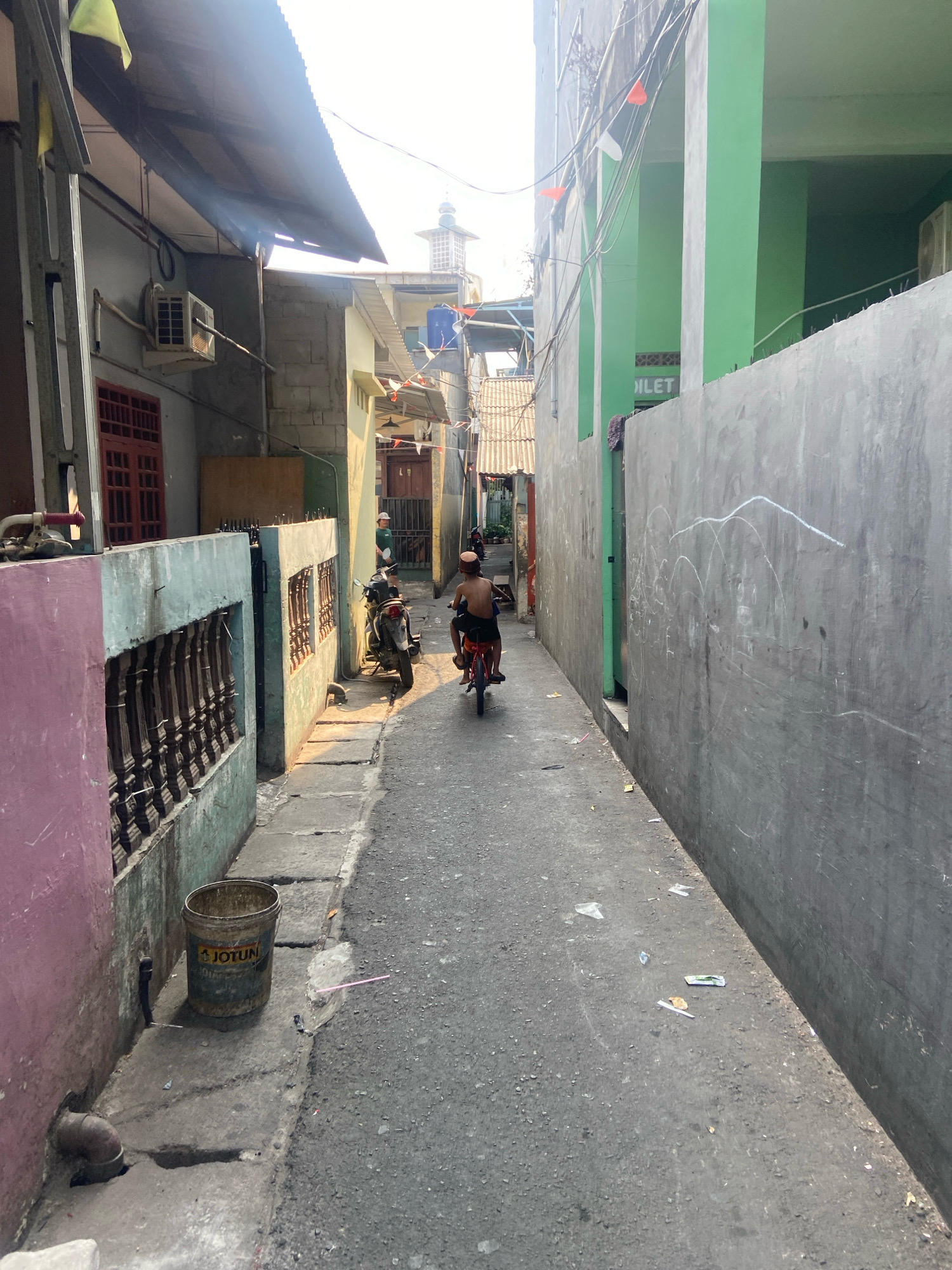 The image depicts a narrow, paved alleyway in a densely populated urban area of North Jakarta, Indonesia, bathed in the warm light of what appears to be late afternoon. The alley is flanked by buildings of varying heights and colors, suggesting a mixed residential and commercial environment.
On the left side of the alley, a building with a pink and pale blue facade is prominent. Its lower section features decorative wrought-iron bars set into a textured wall. Above this, a corrugated metal awning extends outwards, providing shade. An air conditioning unit is visible mounted on the exterior wall of a building further back. Red-framed windows and doors are also present on this side. A weathered, yellow bucket with the "JOTUN" logo sits on the concrete walkway near the foreground, next to a drainpipe.
The right side of the alley is dominated by a tall, grey concrete wall. White chalk drawings, abstract in nature, are scrawled across its surface. To the right, a building painted in a vibrant green is visible, with a sign that reads "TOILET" in white lettering. Small red and white triangular flags are strung along the top of the green building and a grey wall.
In the middle ground of the alley, a person on a red motorcycle is moving away from the viewer. The rider is wearing orange shorts and a dark shirt, with a light-colored cap on their head. Another motorcycle is parked on the left side of the alley, next to a person in a white helmet and light-colored shirt. The ground of the alley is asphalt, showing signs of wear and tear, with scattered debris, including paper and plastic wrappers. In the distance, a mosque minaret with a patterned top is visible above the buildings. The overall impression is one of a working-class neighborhood with a bustling, lived-in atmosphere.
