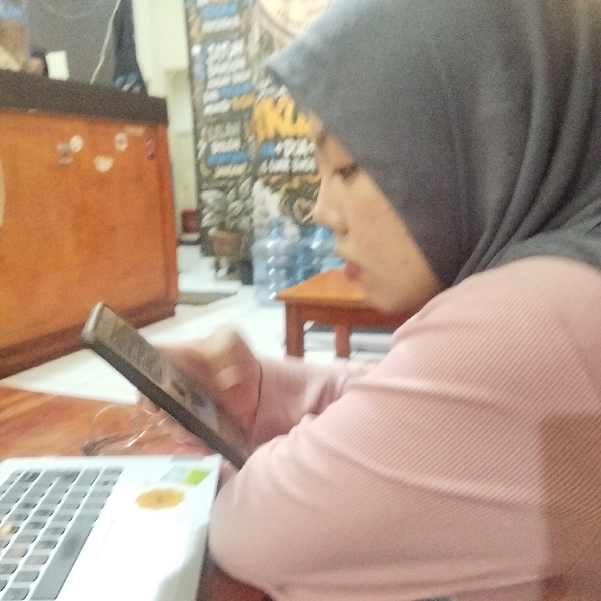 The image depicts a person wearing a grey hijab and a pink, ribbed long-sleeved shirt. They are seated at a wooden table, with a laptop in front of them and holding a tablet device. The person's face is partially visible, in profile, and they appear to be looking at the tablet. In the background, there's an orange cabinet and a wall with posters or decorations. The scene appears to be an indoor setting, possibly a home or a small office, in Sukamanah, Indonesia. The lighting is somewhat dim, suggesting it might be evening or an indoor space with artificial lighting.