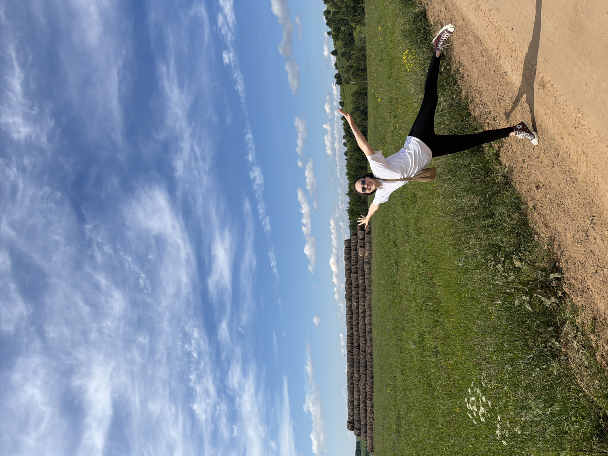 A person is captured in a dynamic pose outdoors on a sunny day in what appears to be a rural or agricultural setting in Van'ki, Russia. The individual, dressed in a white t-shirt, black leggings, and high-top sneakers (possibly maroon or dark brown), is balancing on one leg with arms outstretched and the other leg extended high behind them. They wear sunglasses, and part of their long, light-colored hair is visible over their shoulder, suggesting a joyful or playful moment.
The person stands on a light brown dirt path that occupies the right side of the frame. To the left of the path, a vibrant green field stretches out, dotted with patches of small yellow and white wildflowers. Along the distant edge of this field, a long, low stack of dark brown, cylindrical hay bales forms a distinct line, extending into the background. Behind the field and hay bales, a line of green trees marks the horizon. The upper half of the image is dominated by a bright blue sky, adorned with scattered wispy white cirrus clouds and some smaller cumulus clouds. A clear, long shadow of the person is cast on the dirt path, indicating bright sunlight and daytime. The overall scene conveys a sense of freedom and enjoyment of the natural environment. No visible text is present.