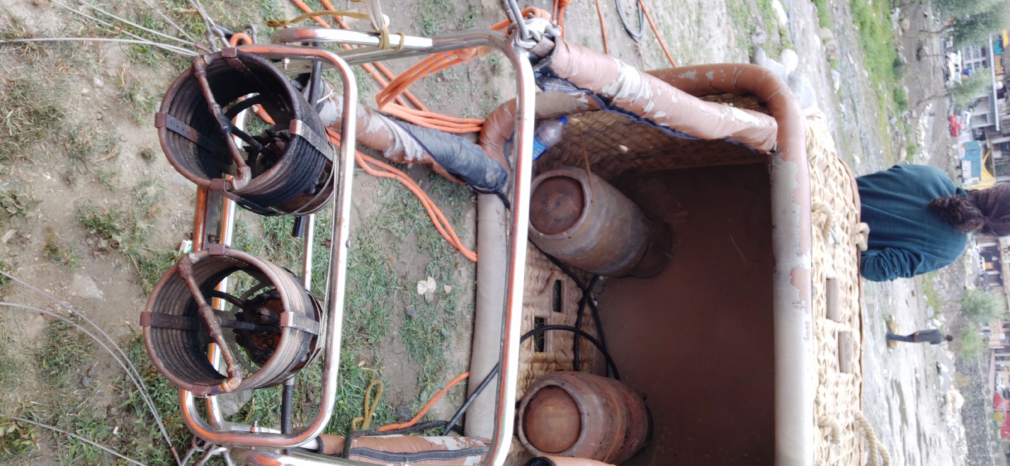 In Kalath, India, a hot air balloon basket sits on the ground, its interior partially visible. Two propane tanks are positioned inside the basket. The exterior of the basket is made of woven material with leather trim, showing signs of wear. Attached to the front of the basket is a metal frame with two burners, ready for operation. Orange hoses are connected to the burners and extend out of frame. A person with dark, curly hair, wearing a dark teal sweatshirt, stands with their back to the camera, looking away from the basket towards a distant hillside. Another individual, dressed in dark clothing, stands further in the background. The ground is a mix of dirt and sparse green grass. The scene suggests preparation for a hot air balloon flight.