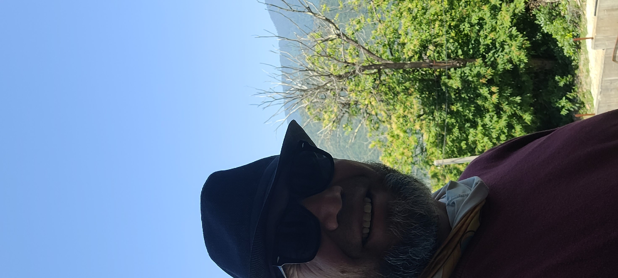 The image shows a close-up, sideways selfie of a person wearing a dark hat and sunglasses. The person has grey facial hair and is smiling with their mouth open, showing teeth. They are wearing a dark red or maroon shirt. The background is a bright blue sky with a blurry tree and greenery behind them. A light-colored scarf or cloth is visible around their neck. The setting appears to be outdoors on a sunny day. The orientation of the photo suggests it was taken by the person themselves, looking up and to the side. The quality of the image is somewhat low, possibly due to the angle or lighting. There is no visible text in the image.