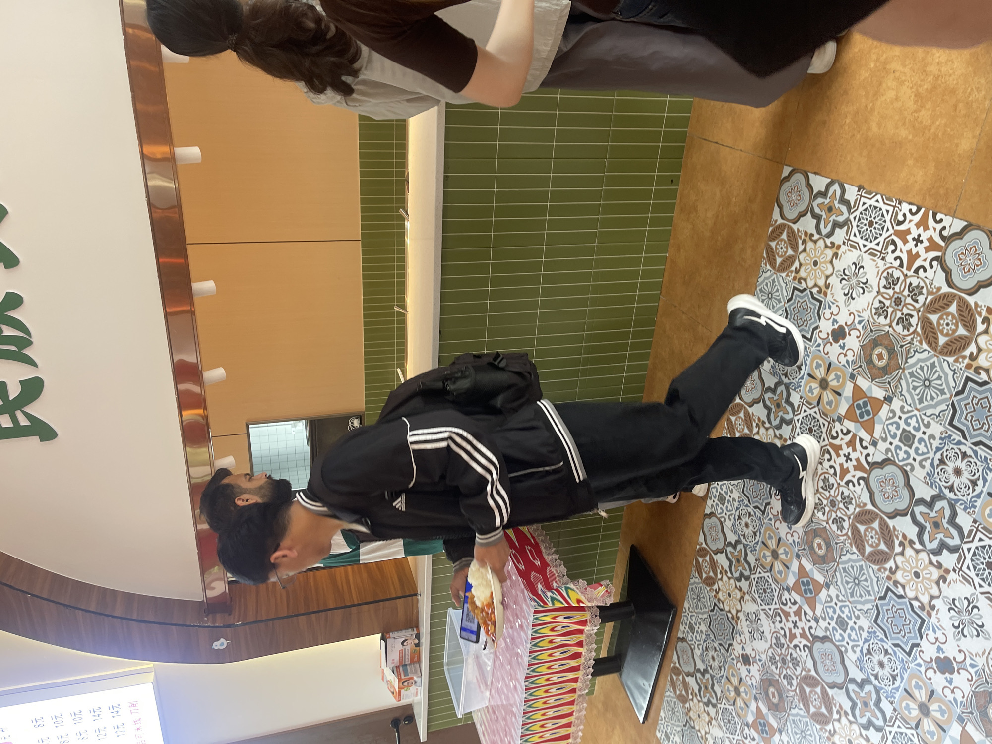 A man wearing black pants and a black jacket with white stripes is standing in front of a display counter holding a plate of food. The man is wearing glasses and has a dark beard. The display counter is decorated with red and white fabric. The floor is tiled with a geometric pattern in various shades of brown, gray, and white. In the background, there is a green tiled wall and a counter with a sign written in Chinese. The image was taken in Chongqing High-tech Zone, China.