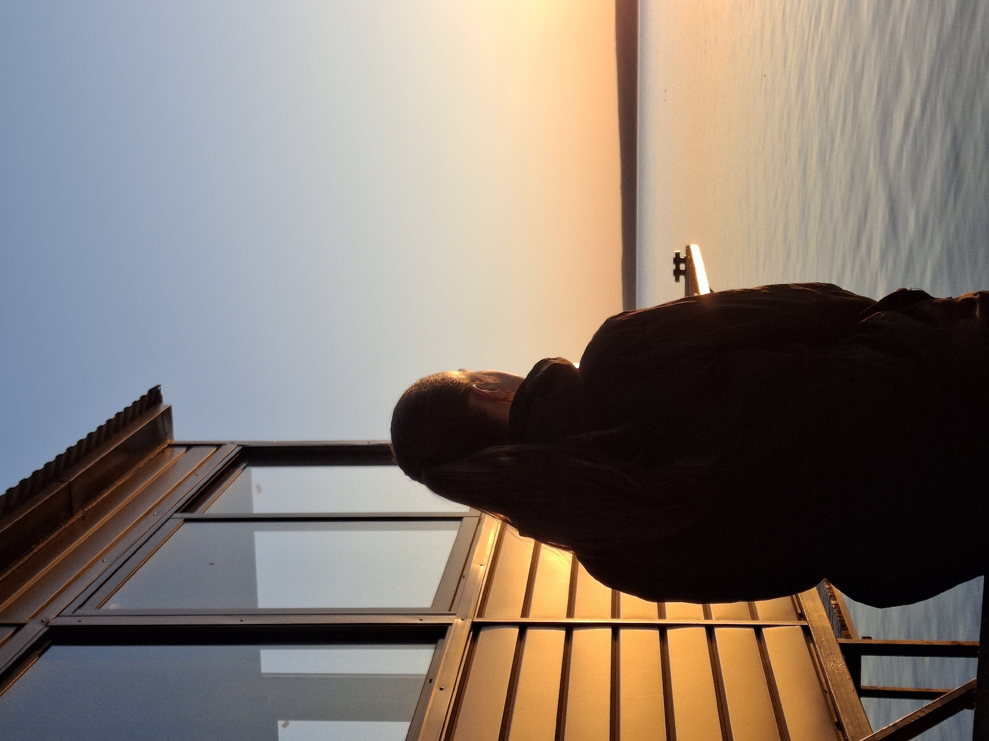 A person is looking out at the sunset over the Kama River from a building in Chaikovsky, Russia. The building has a unique roof made of brown metal corrugated panels and large windows. The person is in silhouette and is standing on an orange metal balcony. The sky is a gradient from blue to orange, indicating the setting sun, and the Kama River is visible in the distance.