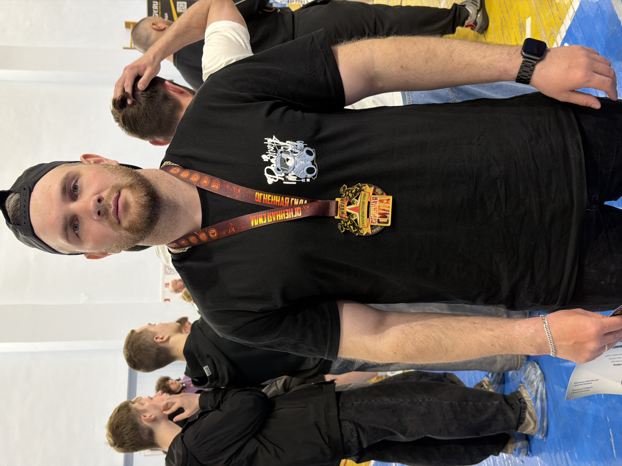 A medium shot captures a man wearing a black t-shirt, black cap, and a medal around his neck. The medal has a red and gold ribbon and features Cyrillic text. The man is fair-skinned with a short beard and is looking directly at the camera. He is wearing a black smartwatch on his left wrist and a silver bracelet on his right wrist. He is standing on a blue and yellow floor.
In the background, there are several other people, mostly men, some wearing black clothing, one in a white shirt. One man is touching the hair of another. The scene appears to be indoors, possibly a gym or event space.
The Cyrillic text on the medal and ribbon suggests a Russian-speaking region, and while not immediately visible in the image, the reference to Izhevsk, Russia, indicates that this is likely the location of the photograph.