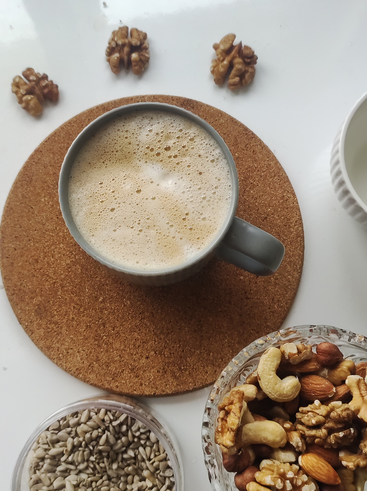 A top-down shot shows a breakfast spread on a white surface, likely a table. In the center, a grey mug filled with frothy coffee sits on a round, brown cork coaster. Several walnuts are scattered around the mug, with three clearly visible. To the right of the mug, a portion of a white ribbed bowl is visible. In the foreground, two clear containers hold snacks. The container on the left is filled with sunflower seeds, while the one on the right is a decorative glass bowl containing a mix of nuts, including walnuts, cashews, almonds, and hazelnuts. The overall scene suggests a simple, healthy breakfast or snack setting. There is no visible text. The lighting appears to be natural, suggesting daytime, possibly morning, and there are no indications of the weather or specific location beyond the provided context. The image evokes a sense of calm and simplicity.