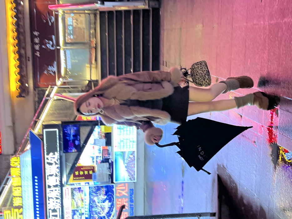 The image depicts a nighttime urban scene in Yuzhong District, China, characterized by artificial light sources and wet pavement. A single female subject is positioned centrally, walking from the background towards the foreground.
The female has long, light brown hair. She is wearing large hoop earrings, a gold-colored necklace, a dark top, a brown bomber-style jacket with significant light brown fur trim on the collar, cuffs, and hem, a black mini-skirt, light grey knitted leg warmers, and brown platform boots. She carries a closed black umbrella in her left hand, which points downwards and features the text "INDI HOT." In her right hand, she holds a small leopard-print handbag. Her gaze is directed towards the viewer's left.
The ground is wet, reflecting the colorful neon lighting. To the subject's left, a structure resembling a boat's bow is illuminated with blue and white neon lights, displaying Chinese characters "游船售票处" (Ferry Ticket Office) and "TICKET OFFICE" in English. Multiple screens show various images, including cityscapes and boats. To the subject's right, a set of dark stairs ascends. Above the stairs, a building entrance is visible, illuminated by orange/yellow lights, with Chinese characters, likely "山城粑粑" (Mountain City Baba), above it. Red neon accents are also present in the background.