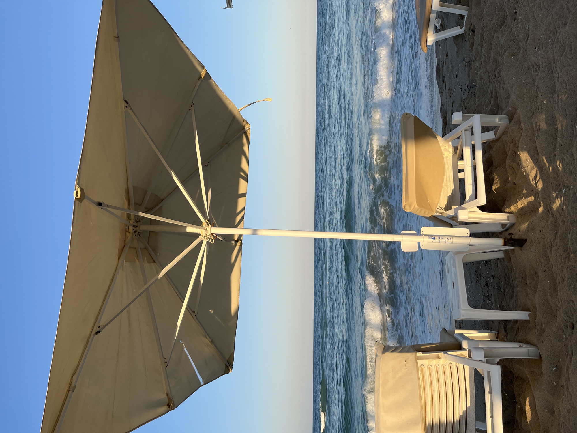 The image depicts a serene beach scene in Obzor, Bulgaria, with the calm turquoise waters of the Black Sea stretching towards the horizon under a clear blue sky. In the foreground, two empty white and tan sun loungers are set up on the sandy beach, partially shaded by a large beige umbrella. A small white table sits between the loungers. Gentle waves are seen lapping at the shore, creating a peaceful ambiance. The overall scene suggests a quiet, relaxing day at the beach.