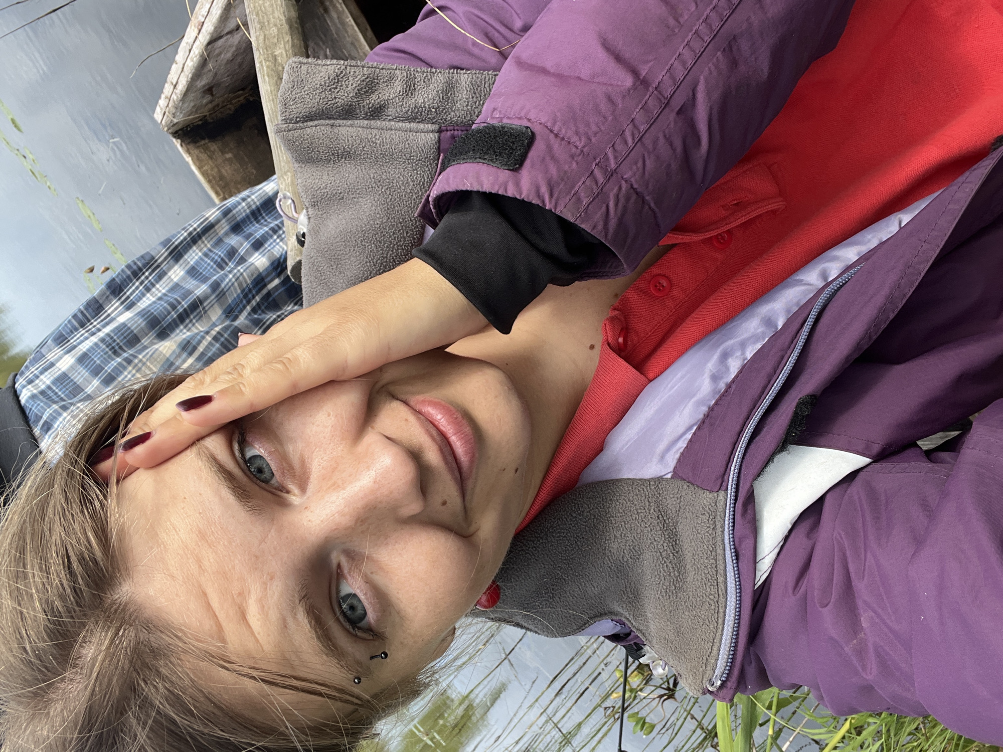 In the image, a young Caucasian woman is taking a selfie while sitting in a wooden boat on a body of water. She has light brown hair, blue eyes, and is wearing a purple jacket over a red polo shirt. She has a piercing above her left eyebrow and is wearing red earrings. Her right hand is touching her forehead. The background shows water with lily pads and reeds, and the edge of the boat, suggesting an outdoor, natural setting. The location is identified as Навасёлкаўскі сельскі Савет, Belarus.