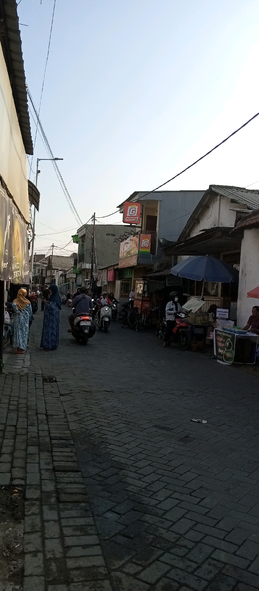 This is a ground-level, full-shot photograph taken in Wadungasri, Indonesia, looking down a street lined with buildings and storefronts. The street is paved with interlocking brick pavers. On the left side of the street, two women wearing hijabs stand on a raised sidewalk. The woman on the left wears a patterned blue and white dress and a yellow hijab, while the woman on the right wears a solid dark blue dress and a matching hijab. Behind them, a building has a large banner with text, partially obscured. Further down the street, several people on motorcycles are moving in the same direction as the camera.
On the right side of the street, various businesses are visible. There is a blue umbrella providing shade over a stall with items displayed. A sign with a red "G" logo and text "GEPREKIN" is prominently displayed on one of the buildings. Another stall sells food, with items like "NASI KUNING" visible on a sign. A man in a dark shirt is seated near this food stall. The sky is clear and pale blue, indicating it is likely daytime. The overall scene depicts a busy street with commercial activity and pedestrian and vehicular traffic.