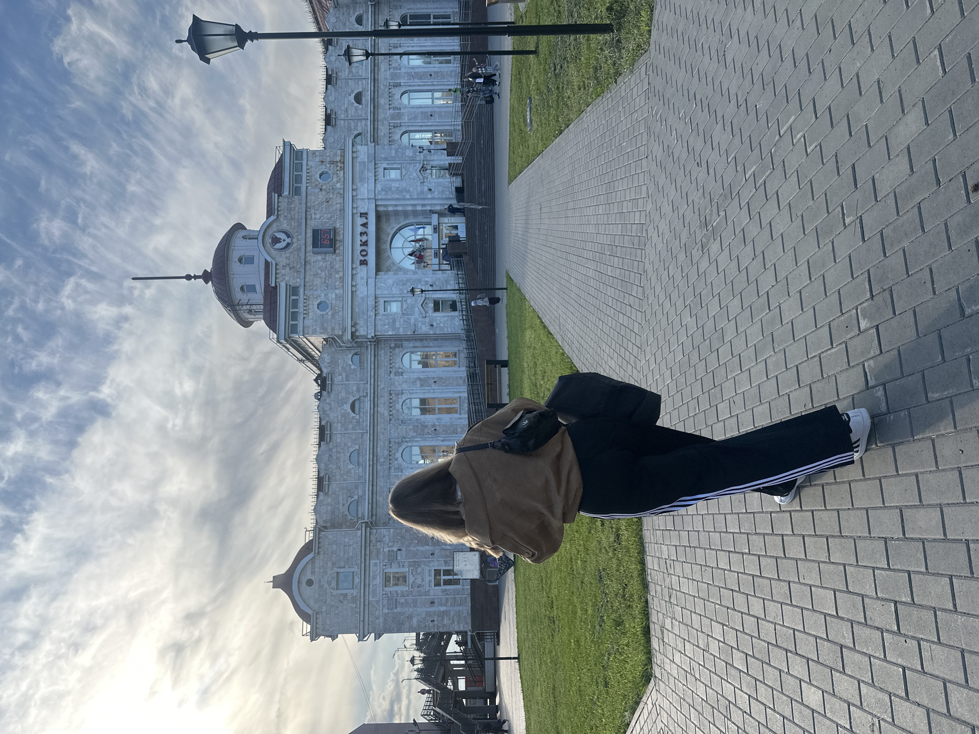 The image captures an outdoor scene in Izhevsk, Russia, featuring a prominent architectural structure, likely a train station, under a bright, partly cloudy sky. In the foreground, a person is seen from behind, standing or walking on a gray paved pathway that stretches towards the building. The person wears a light brown jacket, black trousers with white side stripes, and white sneakers, with a black crossbody bag slung over their shoulder.
The main subject is a large, stately building constructed from light-colored stone, characterized by its classical architectural elements, including arched windows, decorative details, and a central dome topped with a spire. The Cyrillic word "ВОКЗАЛ" (VOKZAL), meaning "Station," is prominently displayed in capital letters above the main entrance. A digital clock on the building's facade shows the time as "6:57". Several other smaller figures can be seen near the building's entrance and on its steps in the distance.
To the left of the pathway, a strip of green grass runs parallel to the building. A tall, black street lamp with an ornate design is visible against the sky on the left side of the frame. The sky is clear with scattered, wispy white clouds. The sunlight creates clear shadows on the paved ground, indicating it is daytime, possibly early morning or late afternoon. The overall impression is one of a functional public space with historical architectural significance.