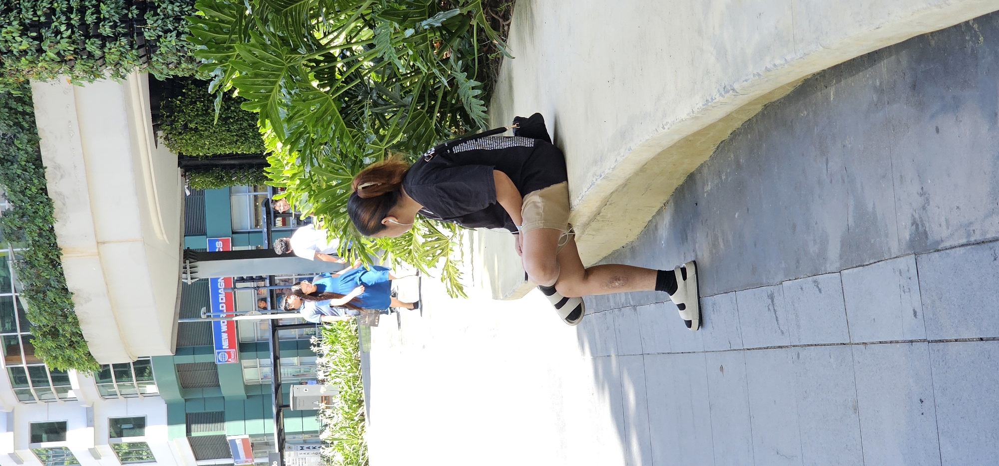 A woman with a brown ponytail, wearing a black t-shirt and khaki shorts, sits on a light-colored concrete bench with her legs crossed. She has on white and black striped socks with white sandals and black socks with white sandals. Her left knee has a noticeable scar. She is wearing white earbuds. A black bag is slung over her right shoulder. She is looking to her left. In the background, there are several people walking, including a woman in a blue dress and a man in a blue shirt. Behind them is a modern building with green and white accents, and a vertical garden on the right side of the image. The scene appears to be an outdoor public space with a tiled walkway and a curved concrete divider. The location is the Ayala Center in Makati City, Philippines, specifically near the Greenbelt 5 shopping mall. The weather is sunny, casting shadows on the ground.