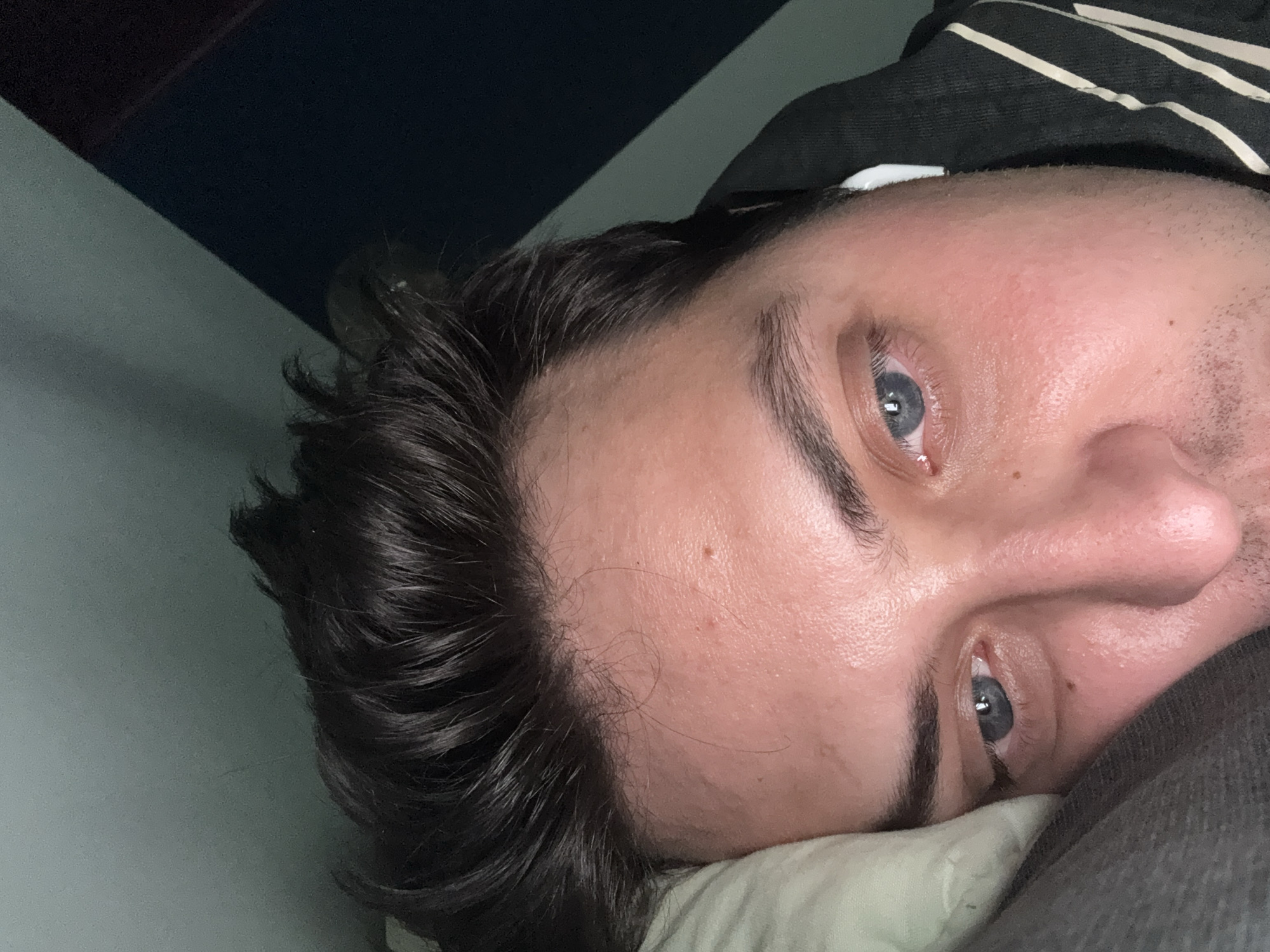 A close-up, angled portrait shows a man lying on a pillow. He has dark hair, blue eyes, and fair skin. He appears to be indoors, possibly in bed, with a wall visible in the background. The lighting is ambient. There is no information in the image to suggest the location is Vidnoye, Russia.