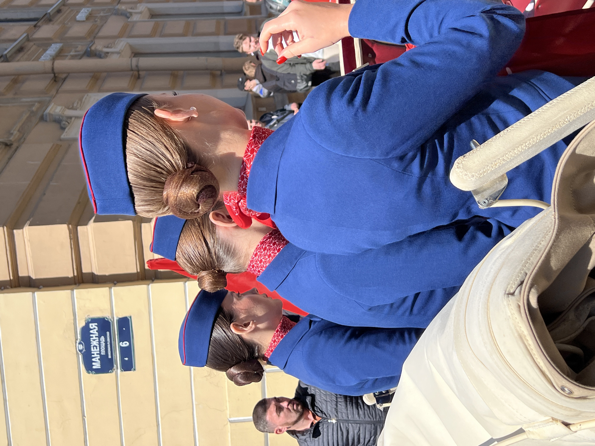 The image shows three women wearing matching blue uniforms with red trim and red patterned scarves, their hair styled in buns. They are positioned in a line, with only the first two being fully visible from the shoulders up. A man is standing below the third woman in the line, facing the same direction. Behind the women, several other blurred figures can be seen in the background. A sign on the building to the left of the women reads "MANEZHNAYA," indicating they are located on Manezhnaya Square in Saint Petersburg, Russia. A white pram partially obscures the view.