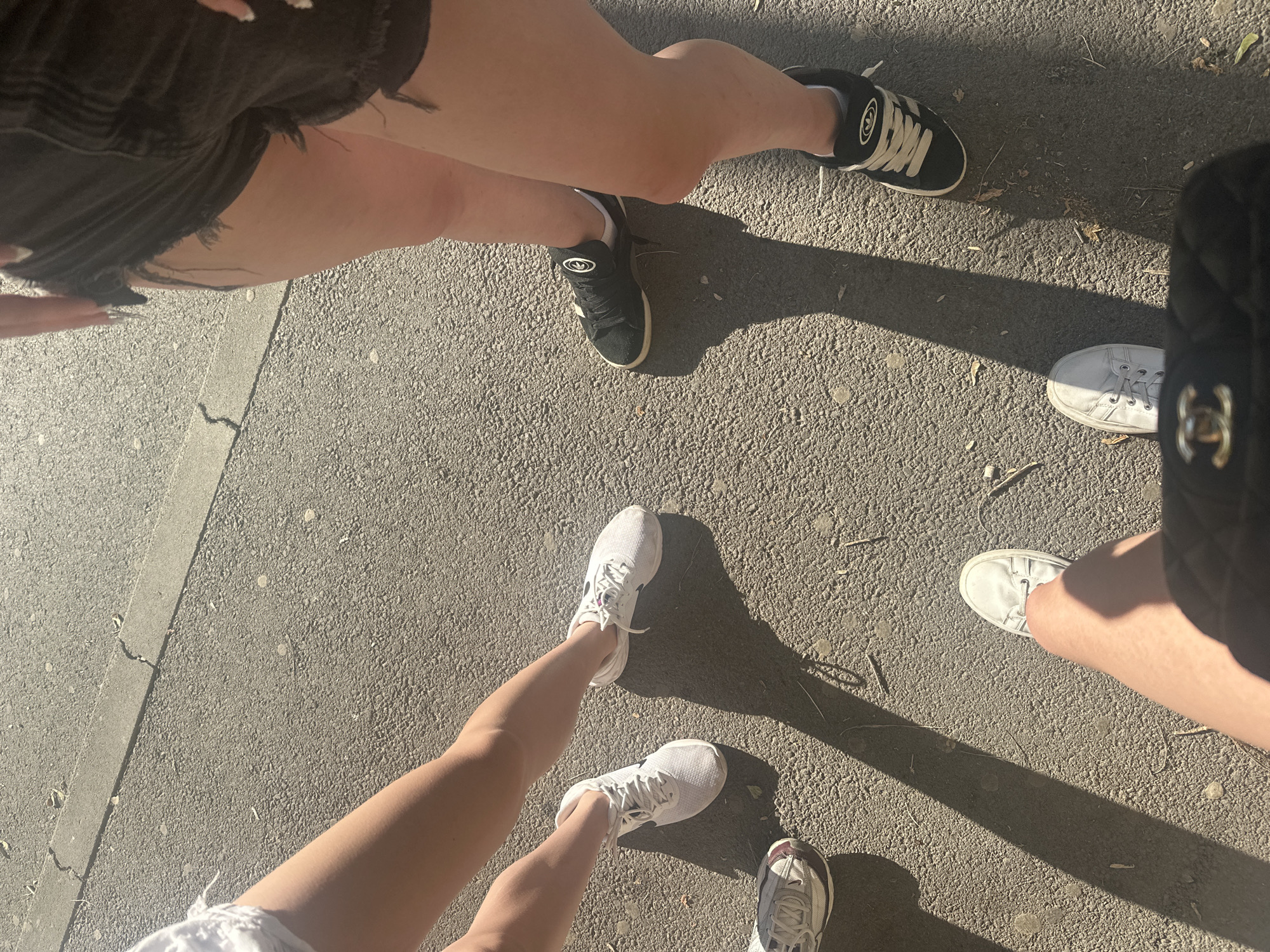 The image shows the lower legs and feet of at least three people standing on a paved surface, possibly a sidewalk or street. One person is wearing white sneakers, another black sneakers with white laces and trim, and a third person is wearing white canvas shoes. A black quilted bag with a gold interlocking C logo is visible in the foreground, obscuring the feet of the person holding it. The scene is outdoors, with shadows indicating sunlight. The location is Shumen, Bulgaria.