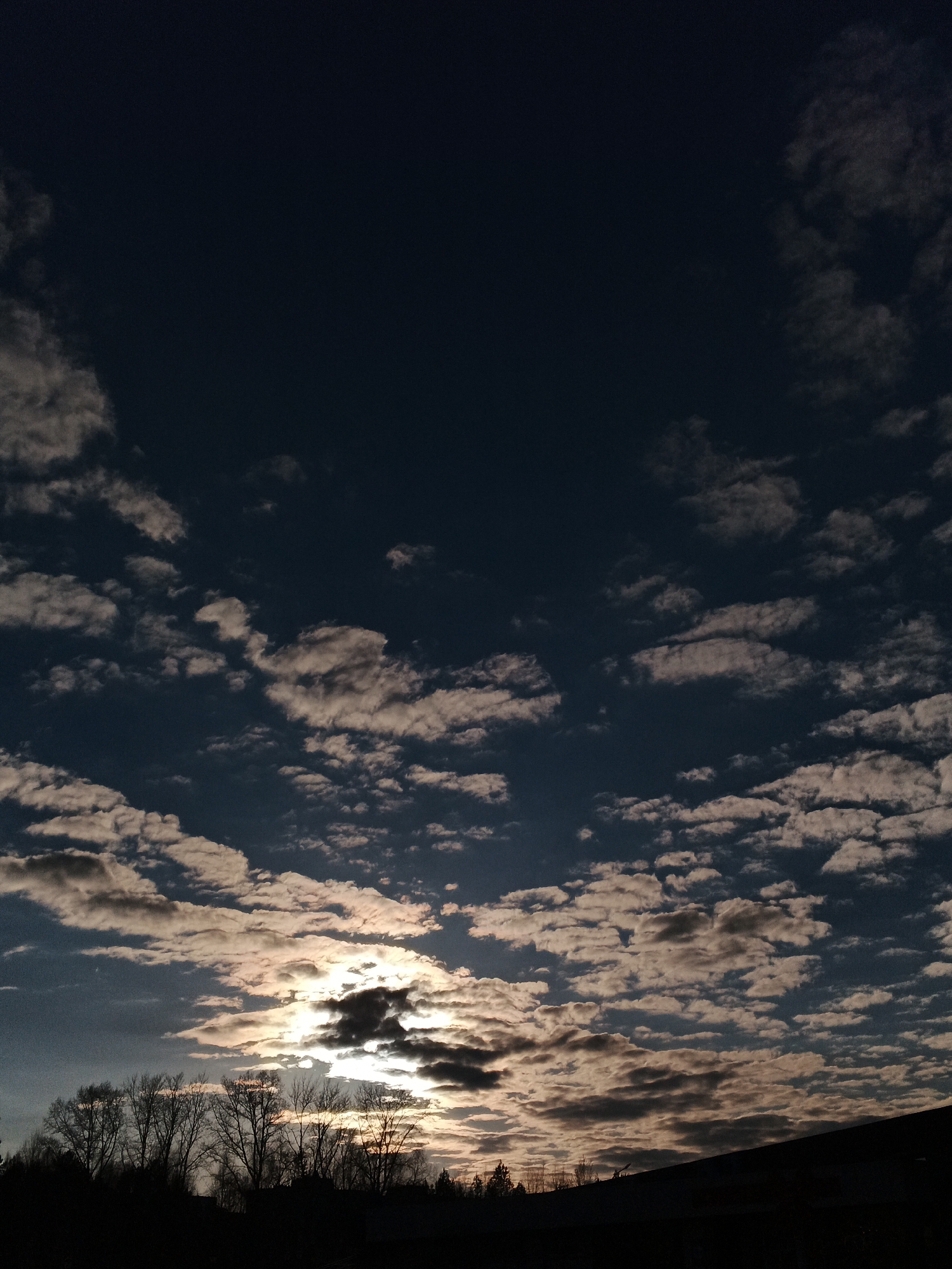 A low-angle shot shows a dark evening sky over what is identified as Chaikovsky, Russia. The sky is predominantly dark blue, filled with small, scattered clouds tinged with light from the setting sun. The sun is partially obscured by clouds near the horizon, casting a bright glow. Below the clouds, silhouetted trees line the horizon, and an indistinct dark shape that appears to be a building. No people are visible.