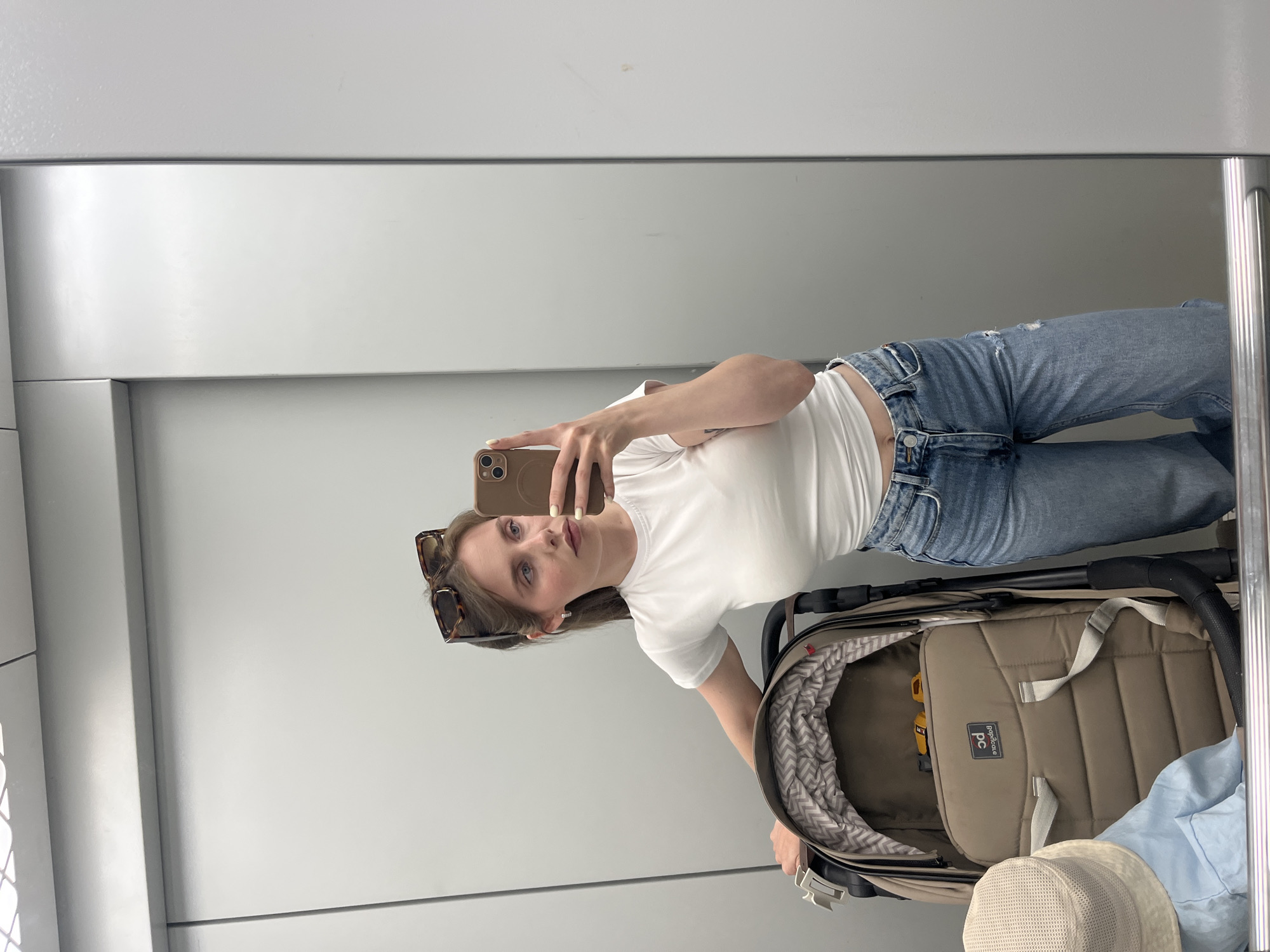 A fair-skinned young woman is seen in a mirror selfie in what appears to be an elevator. She is wearing blue jeans and a white t-shirt. Sunglasses rest on top of her head. In front of her is a beige stroller with a gray and white patterned lining. The stroller appears to contain a light blue blanket and a beige hat. The background is a plain gray, likely the walls of the elevator. I cannot confirm this image was taken in Chaikovsky, Russia.