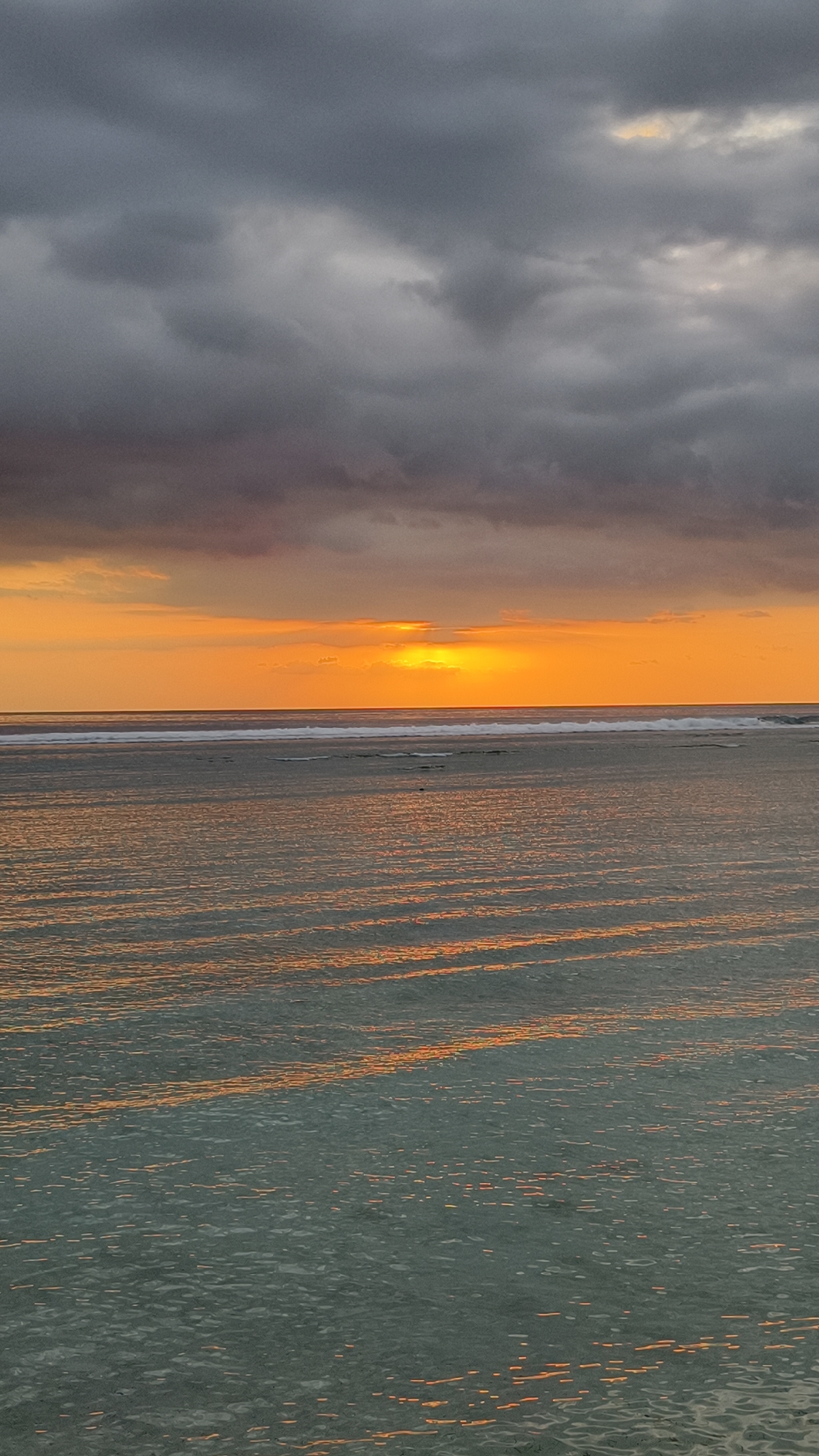 The image depicts a sunset over the ocean. The sky is filled with dark, ominous clouds at the top of the frame, transitioning into a vibrant orange glow near the horizon where the sun is setting. A thin line of waves is visible on the water. The water's surface reflects the orange light, creating a rippled pattern. The location is Unknown, Indonesia.