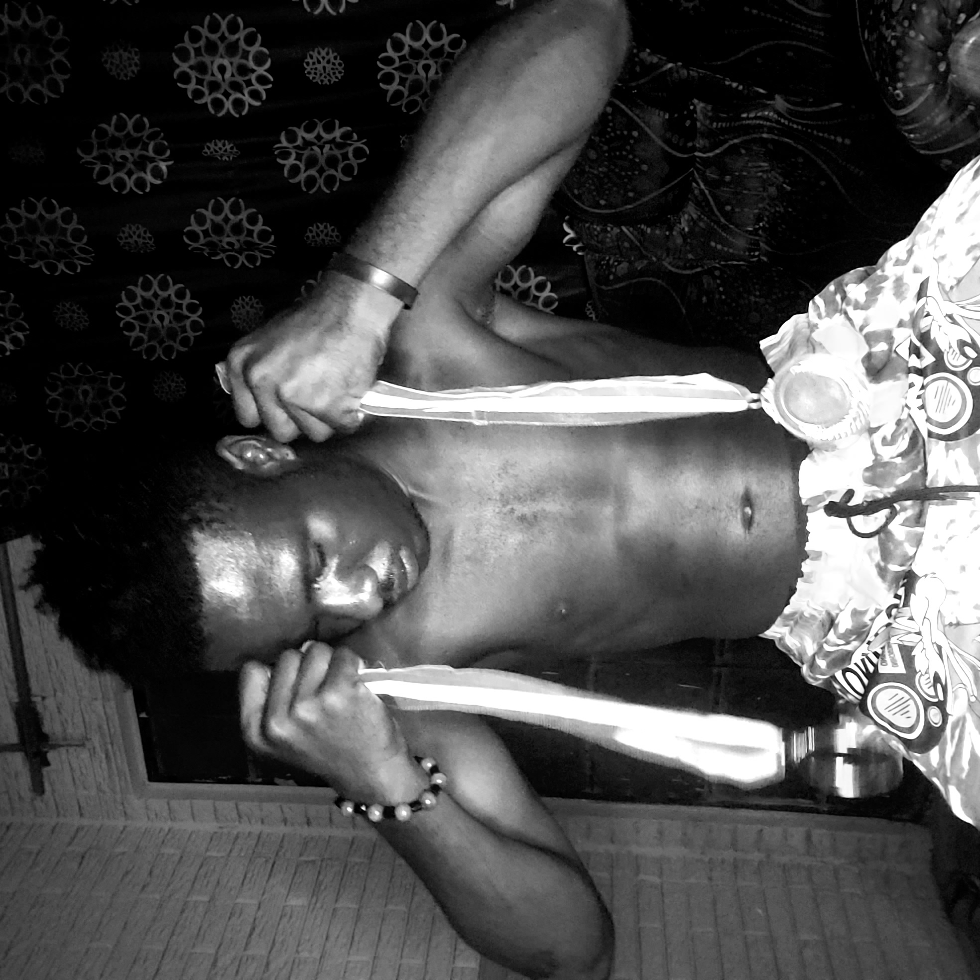 A black and white portrait of a shirtless person with dark hair and brown skin is positioned indoors in Narayi, Nigeria. The person is wearing patterned shorts. The person is holding a white cloth, possibly a towel, to his or her face. One arm has a metallic bracelet and the other a bracelet made of round beads. The background consists of patterned textiles and a wall with a grooved pattern.