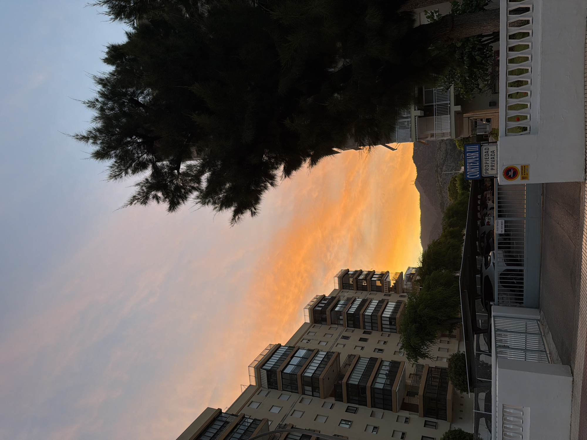 The image depicts a sunset with vibrant orange and pink clouds filling the sky. In the foreground, there are apartment buildings with balconies, a large dark green tree, and a white fence. A sign on the building reads "CORTEMAR III PROPIEDAD PRIVADA". The scene appears to be in a residential area, possibly in a coastal town, given the style of the buildings and the presence of palm trees. A parking area with cars is visible behind a gate. The overall impression is one of a peaceful evening in a Mediterranean-style setting.