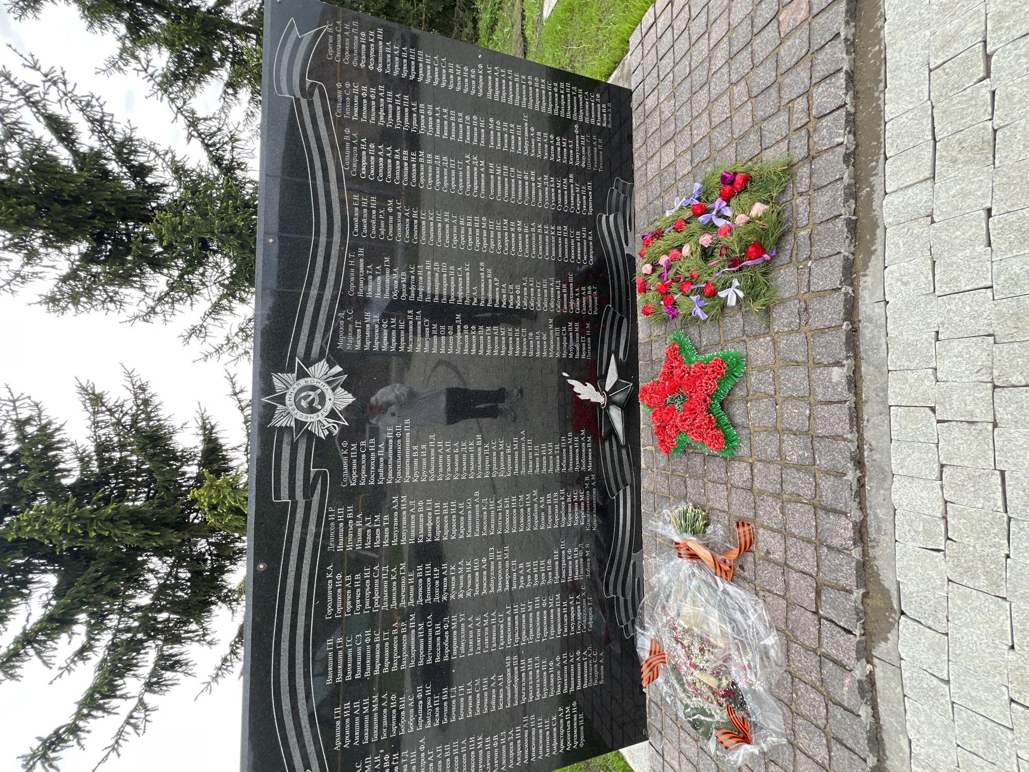 Here is a description of the image:
**Scene:** The image depicts a memorial monument in Введенская Слобода, Russia, likely a war memorial. The memorial is a large, black granite slab, standing vertically. Engraved on the monument are names in Cyrillic script, organized in columns, likely commemorating individuals. The monument is constructed of granite blocks.
**Objects:**
* Three floral wreaths are placed on the cobblestone ground in front of the monument. One wreath is arranged with red and green flowers in the shape of a Soviet star, another is a traditional circular wreath with assorted colorful flowers, and the third is wrapped in cellophane with an orange and black ribbon.
**Environment:**
* The monument is situated outdoors, next to a sidewalk or paved area constructed of grey cobblestones.
* Green foliage from nearby trees is visible in the background, suggesting a park-like setting.
**People and Actions:**
* A reflection of a person taking the photo is visible on the monument's surface. No other individuals are directly visible.