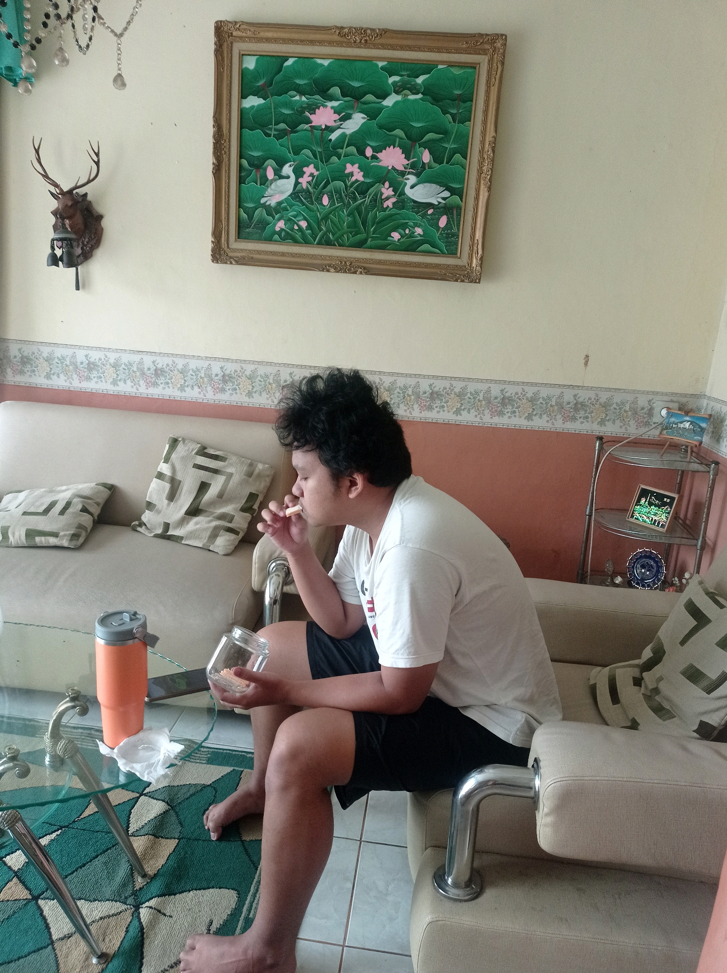The image shows a young man sitting on a couch, smoking. He is wearing a white t-shirt and black shorts. He is holding a glass jar in his left hand and a cigarette in his right hand. There are two pillows on the couch behind him, and an orange thermos on a glass table in front of him. The room appears to be a living room, with a painting on the wall above the couch. The painting depicts a scene with lotus flowers and birds. A deer head decoration is also visible on the wall. A shelf unit with various items is in the background. The overall impression is a casual, indoor setting. The location cannot be determined from the image.