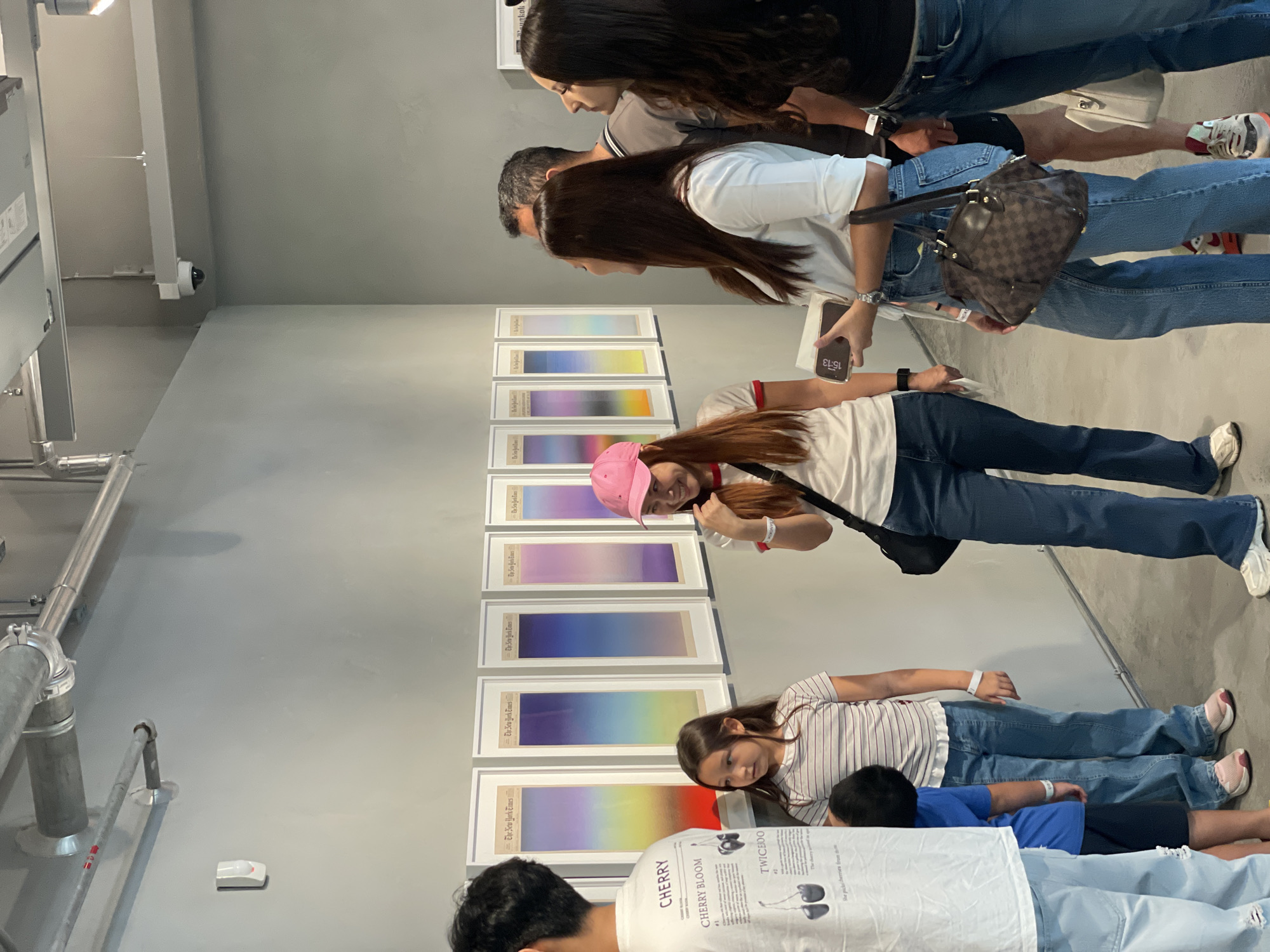 A group of people are viewing artwork in an indoor gallery. The artwork is arranged in a vertical line on the wall, and consists of framed pieces displaying gradients of color. Some people are standing, while others are lying on the floor to view the art. One person is wearing a pink hat. The clothing worn by the people consists of jeans, t-shirts, and shorts. One woman is holding a purse and a cellphone. There are pipes attached to the wall on the left side of the image. Based on the woman's cellphone screen, the location is in Bangkok, Thailand.