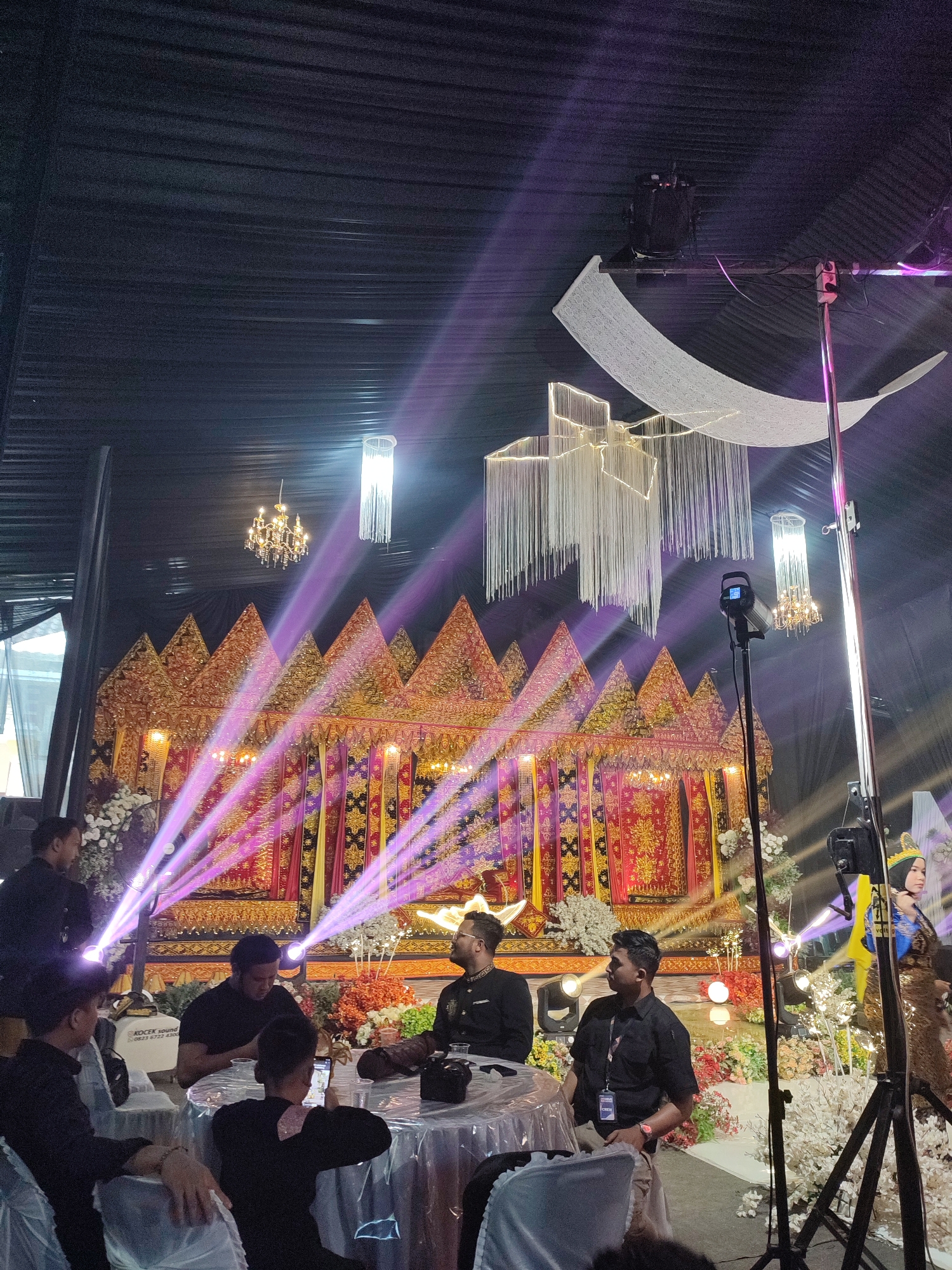 This image captures an indoor event in Muliengmanyang, Indonesia, featuring an elaborately decorated stage or backdrop. The ceiling is draped with dark fabric, from which multiple chandeliers hang, including a traditional golden chandelier, a modern white fringed chandelier, and another white chandelier with long fringes. A large white fabric decoration, resembling a crescent moon, is also suspended from the ceiling.
The stage is adorned with traditional Indonesian *pelaminan*-style decorations, characterized by multiple tiered, pointed structures in gold, red, and purple patterns. Several purple spotlights illuminate the stage from the foreground. Floral arrangements in shades of white, red, orange, and green are visible around the stage and on tables.
In the foreground and midground, approximately 8-10 men are present. Most are dressed in black shirts. Several men are seated around a round table draped with a white or silver tablecloth; some are looking at mobile phones, while others appear to be conversing. A man in a black shirt with a lanyard is standing near a table. Professional photography or videography lighting equipment on a tripod is positioned in the right foreground, and another is visible further back. A white equipment bag or speaker labeled "KOCEK SOUND" with a phone number is situated behind the seated men. Partially visible on the far right, near the stage, is a person (likely a woman) in traditional attire, including a golden-brown patterned outfit, a yellow sash, and a green and gold headpiece.