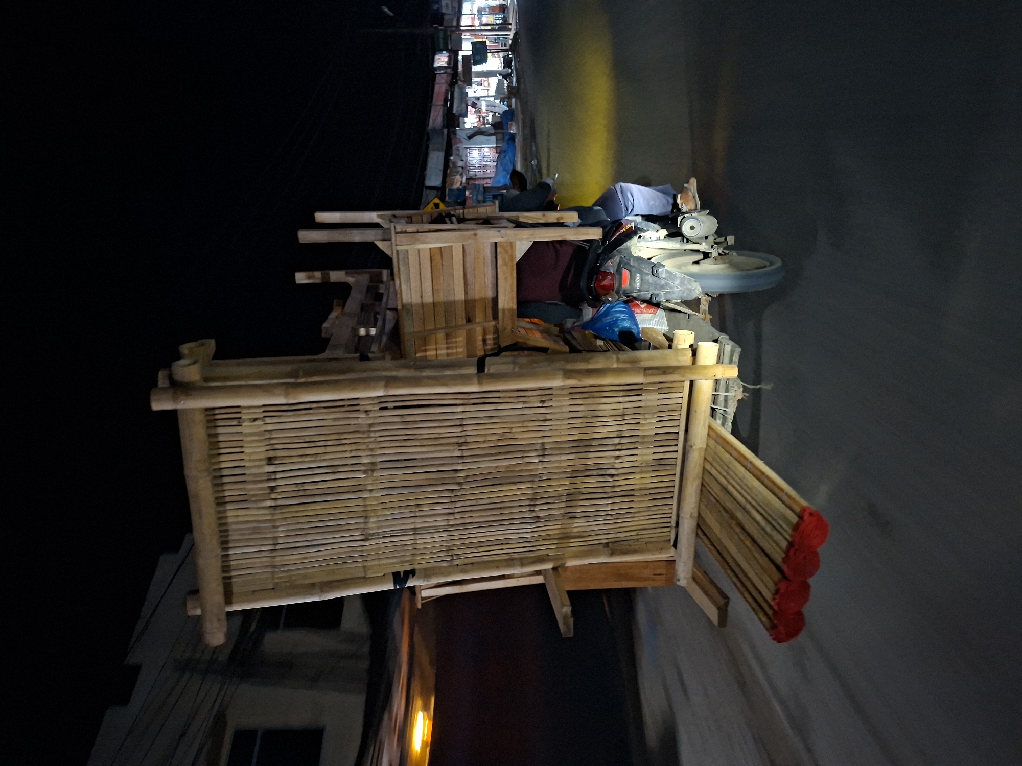 The image, taken at night in Keudegeudong, Indonesia, features a makeshift outdoor workshop. The workshop consists of a bamboo structure, possibly a partially constructed building or a vendor's stall. A man lies on top of the structure, resting near a circular saw and a Honda motorcycle. Adjacent to the bamboo structure, there is a residential area with multiple levels, and electrical wiring is visible. The overall lighting is dim, and the surrounding environment appears cluttered and dimly lit, typical of a busy urban space.