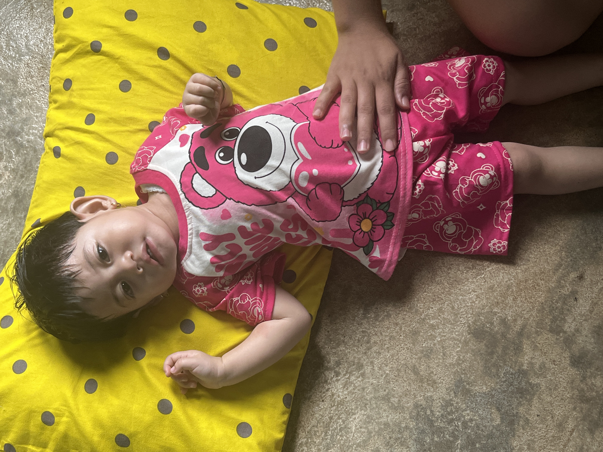 A young child lies on a yellow polka-dotted pillow on a textured concrete floor. The child is wearing a pink t-shirt with a white chest panel featuring a cartoon bear holding a heart, and matching pink shorts with a pattern of bears and flowers. The child's left arm is extended, and a hand is resting on the child's stomach. The background is out of focus, but appears to be an indoor setting. The image was taken in Meliau Hulu, Indonesia.