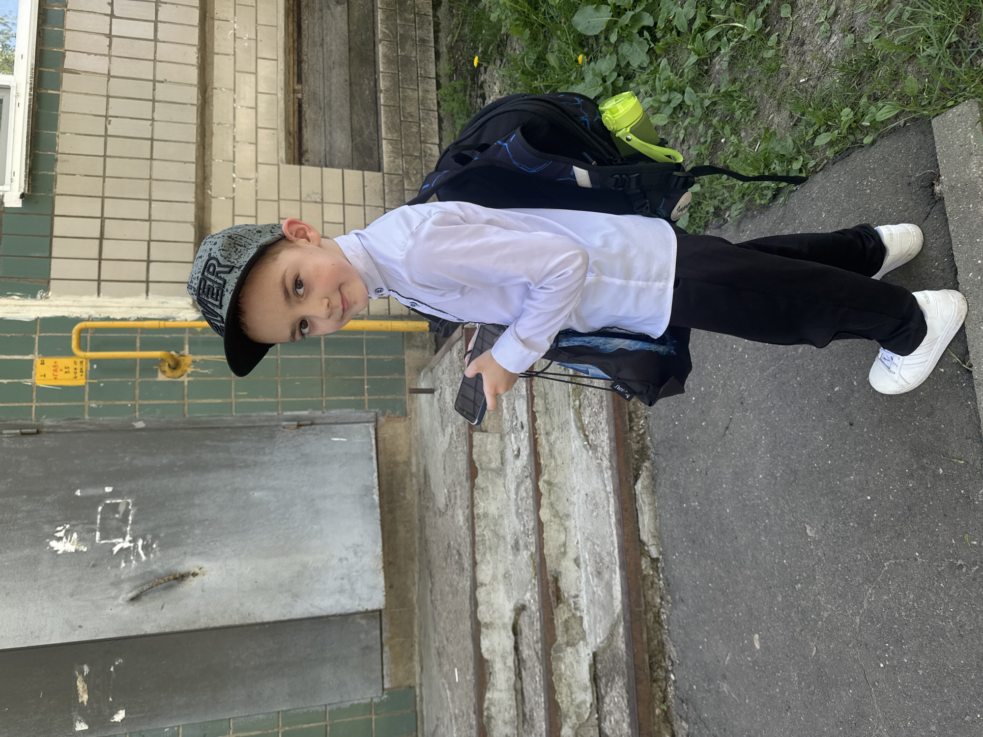 A boy wearing a white long-sleeved shirt, black pants, and white sneakers stands outdoors next to a concrete staircase and a green tiled wall. He's wearing a gray and black baseball cap and holding a black smartphone. A black backpack with blue accents and a neon green water bottle sits on the steps behind him. The ground is paved with gray asphalt. The location is Razdolye, Russia.