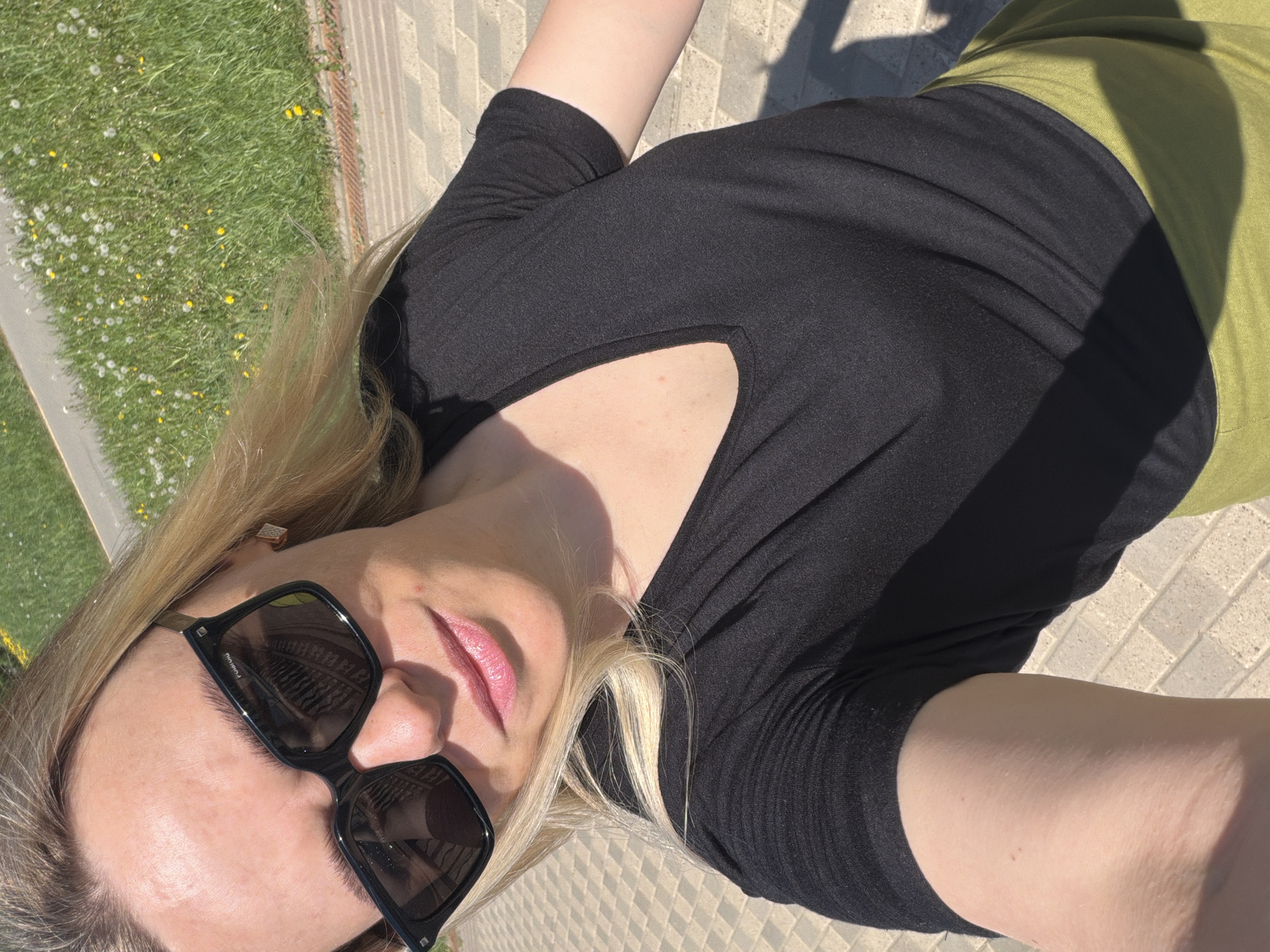 A woman is depicted in an eye-level shot on a sunny day. She is wearing black sunglasses, a black short-sleeved shirt, and a lime green garment. The background includes grass and pavement. The location is Votkinsk, Russia.
